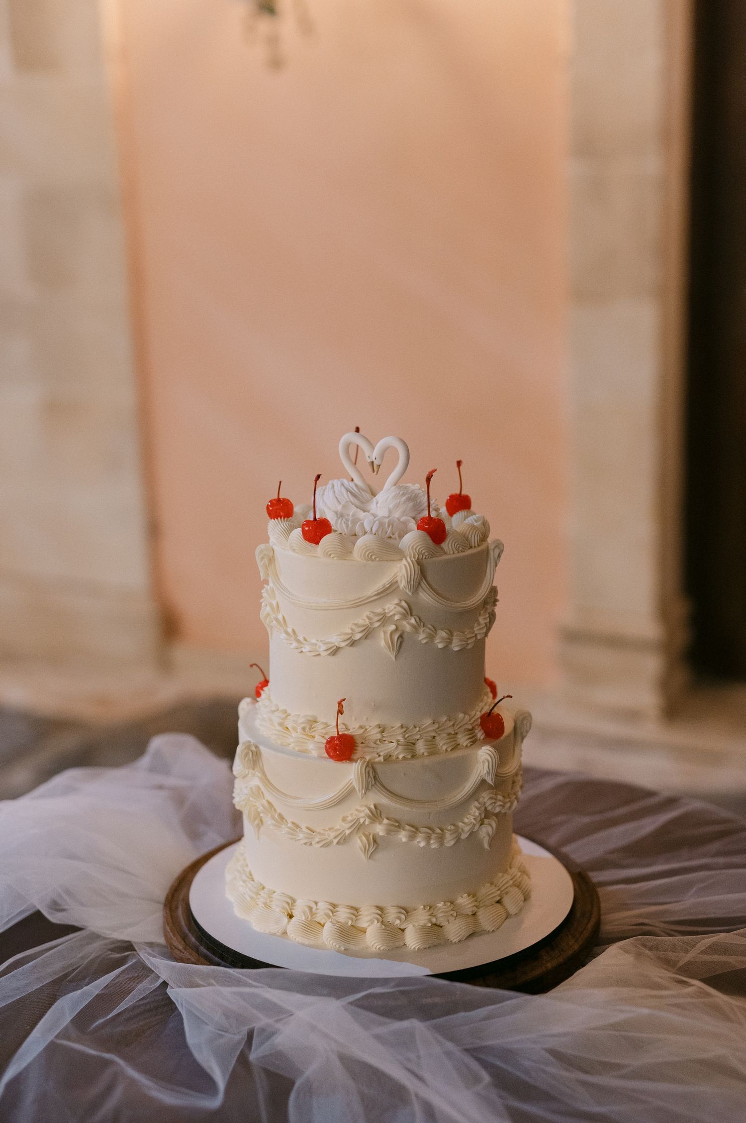 An elegant white three-tiered wedding cake decorated with delicate swirls, red cherries and a swan topper on white tulle.