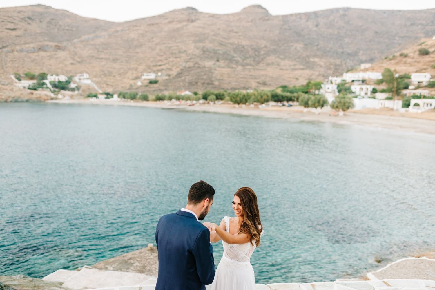 Wedding couple overlooks Mediterranean bay with coastal village in background.
