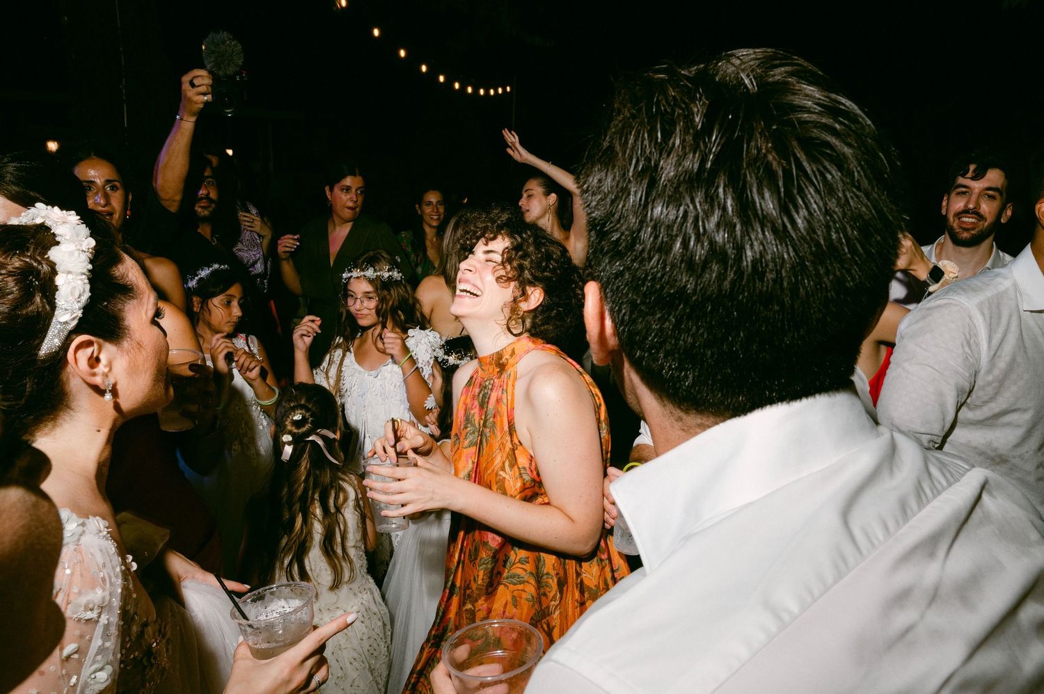 People dancing and celebrating at a lively wedding reception with string lights overhead.