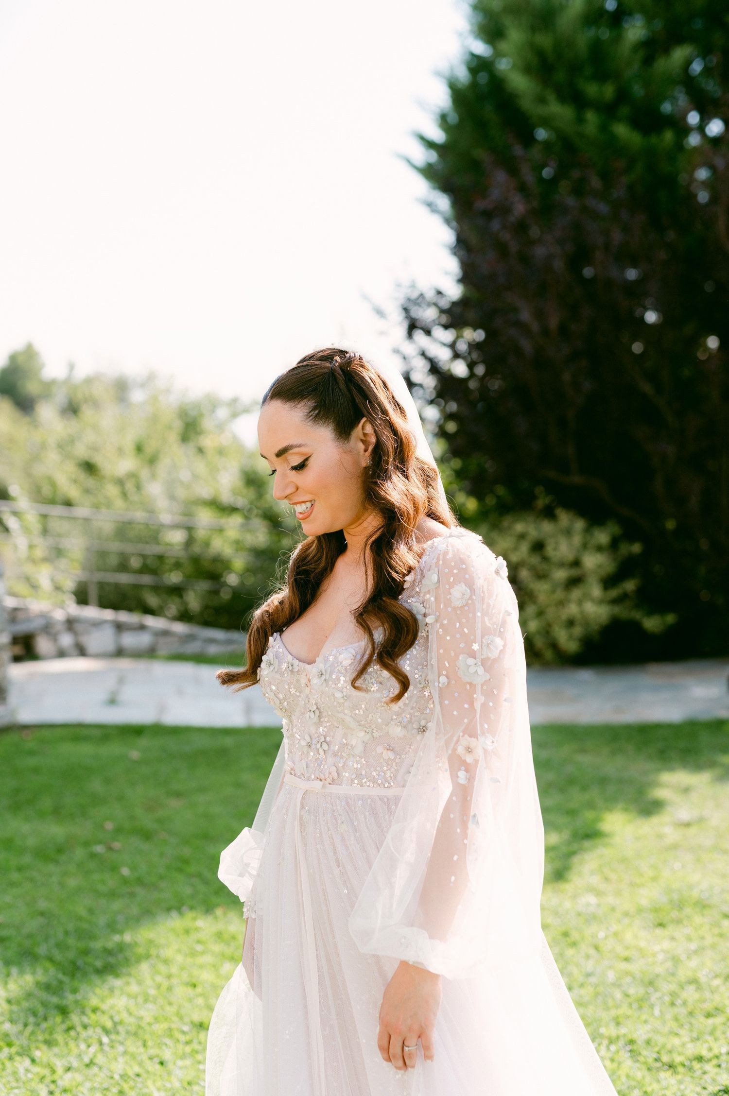 Beautiful bride wearing flowing lace long sleeve wedding gown stands in sunlit garden setting on her wedding day.