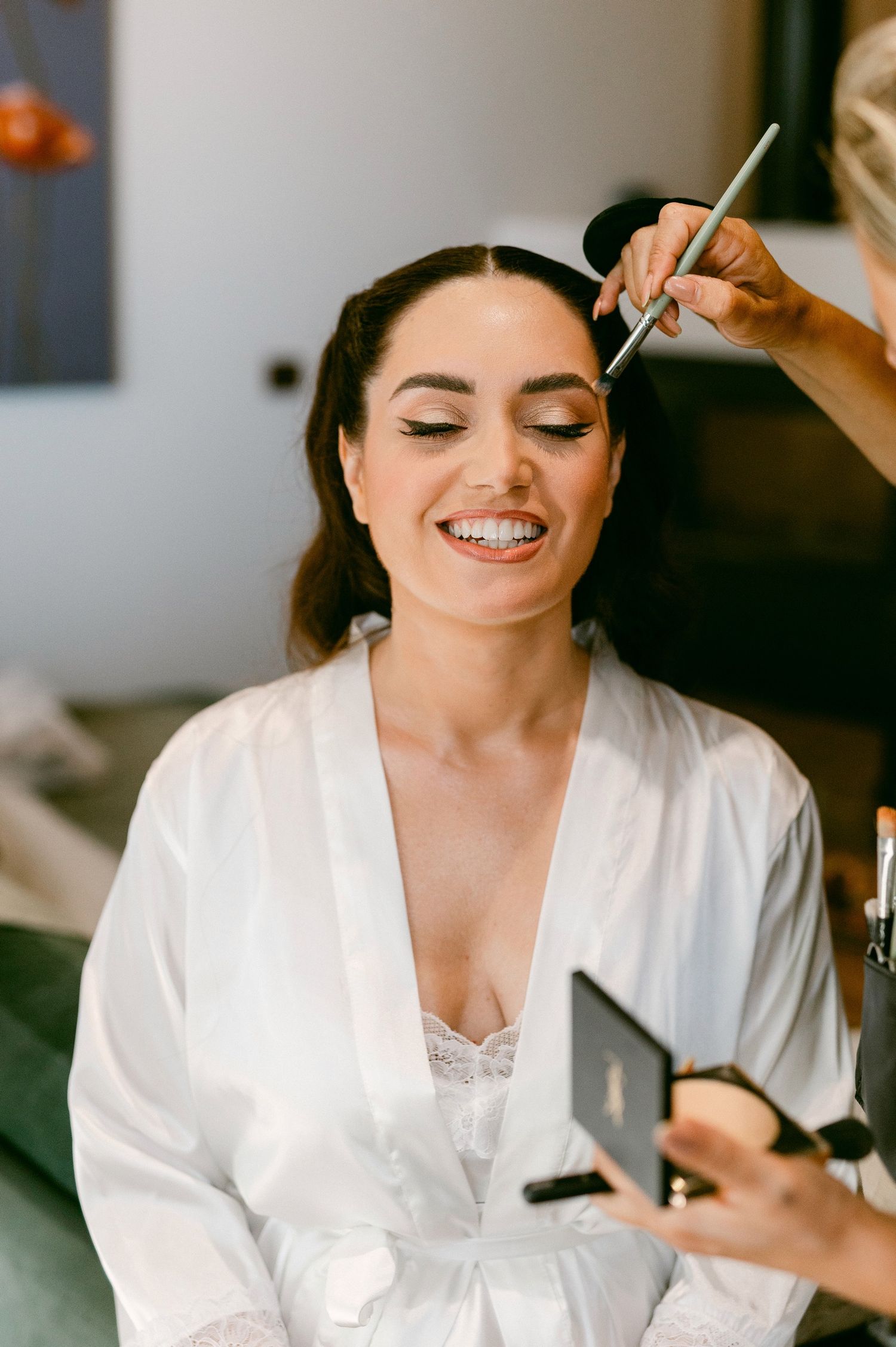 A person in a white robe sits with closed eyes and a smile while having makeup applied with a brush.