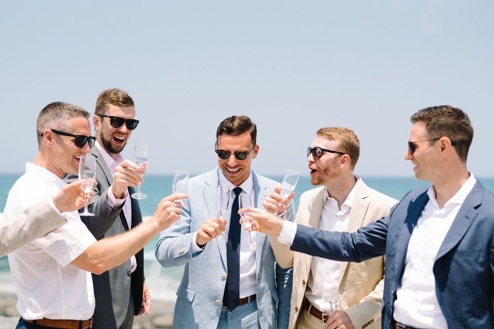Groomsmen in navy suits and sunglasses toast with drinks during outdoor wedding celebration.