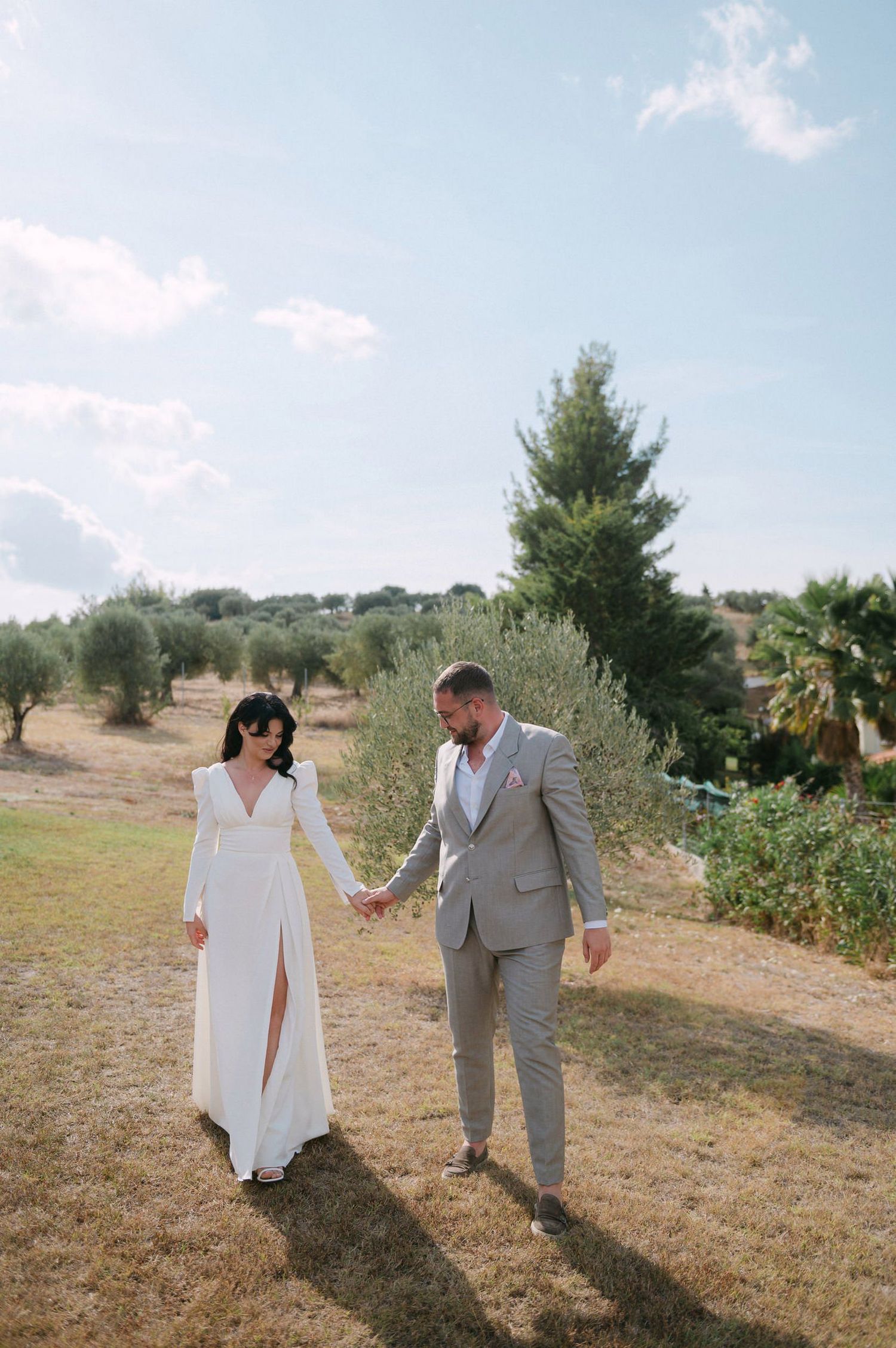 A couple walks hand in hand through a sun-drenched field during their wedding photoshoot.