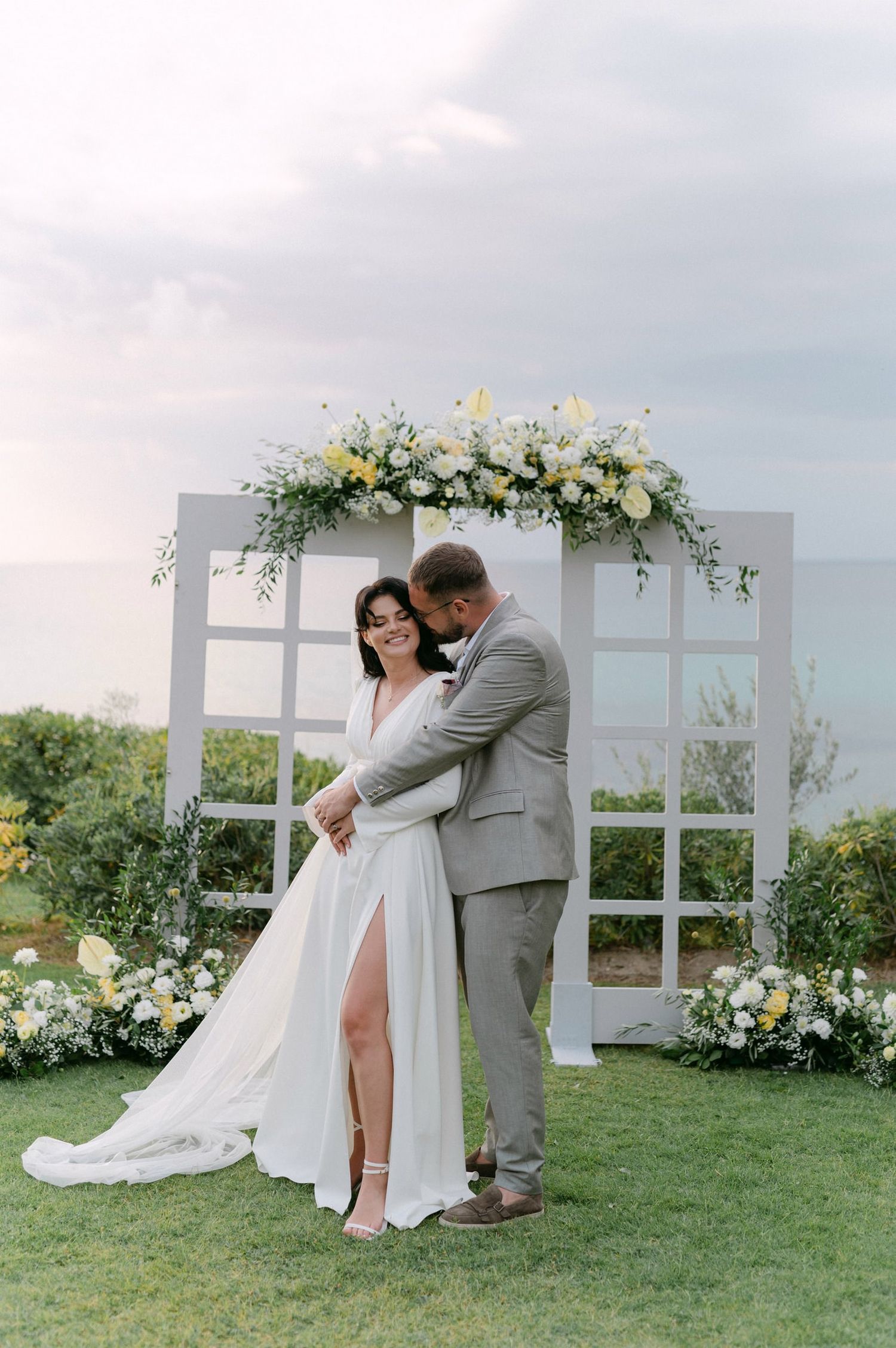 A romantic wedding couple shares intimate moments under a white floral arch in an outdoor garden setting.