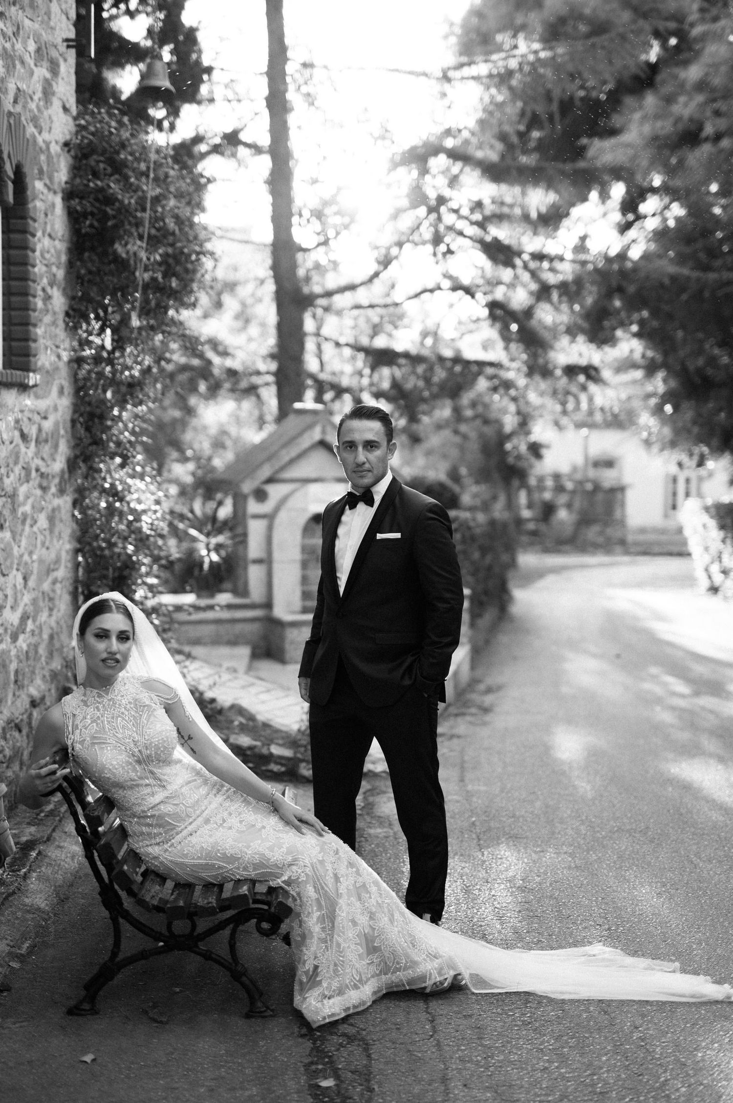 A romantic black and white photo of a couple posing on a park bench surrounded by trees.