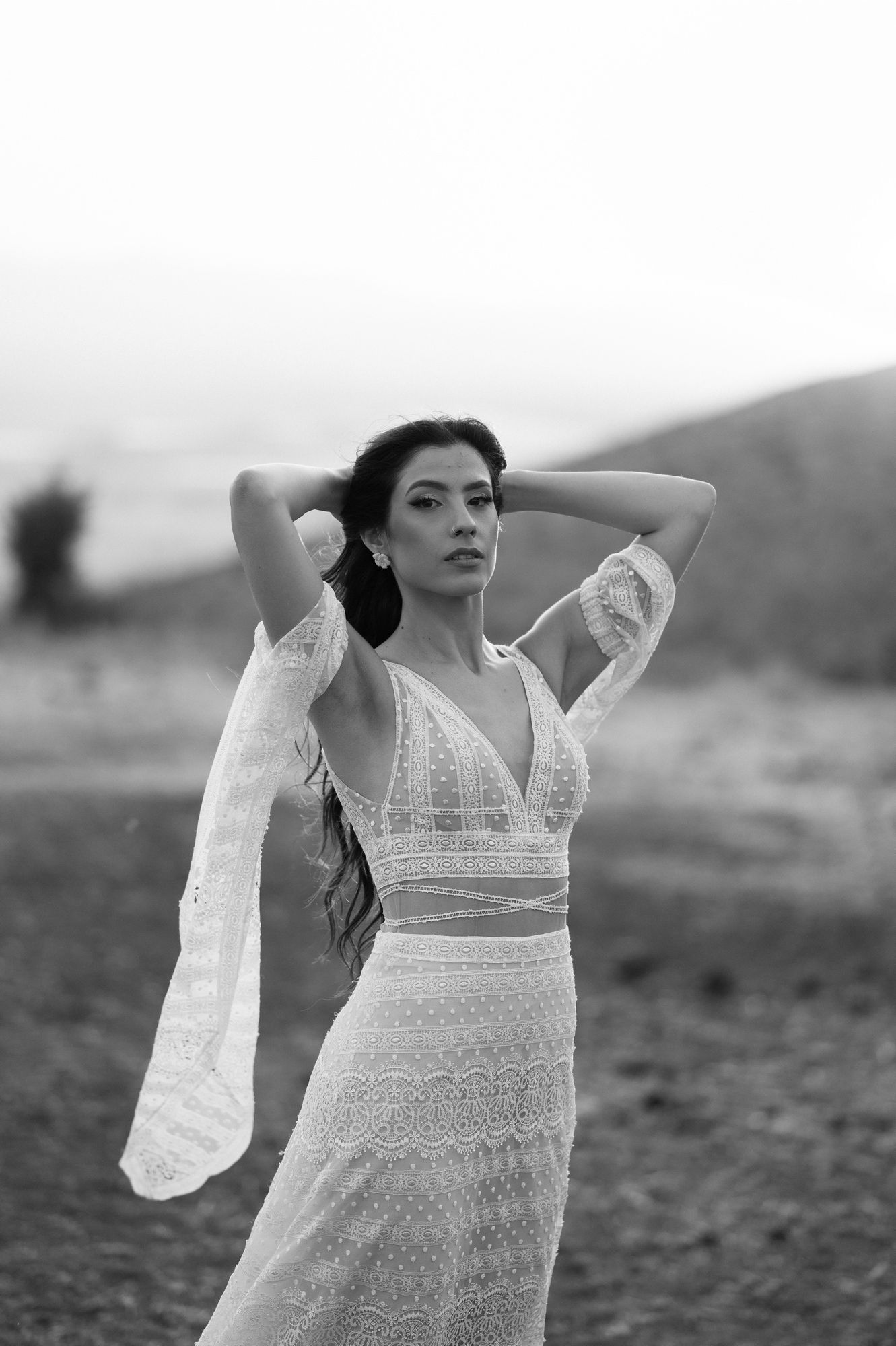 A black and white photograph of someone posing in a delicate white lace dress outdoors with mountains in the background.