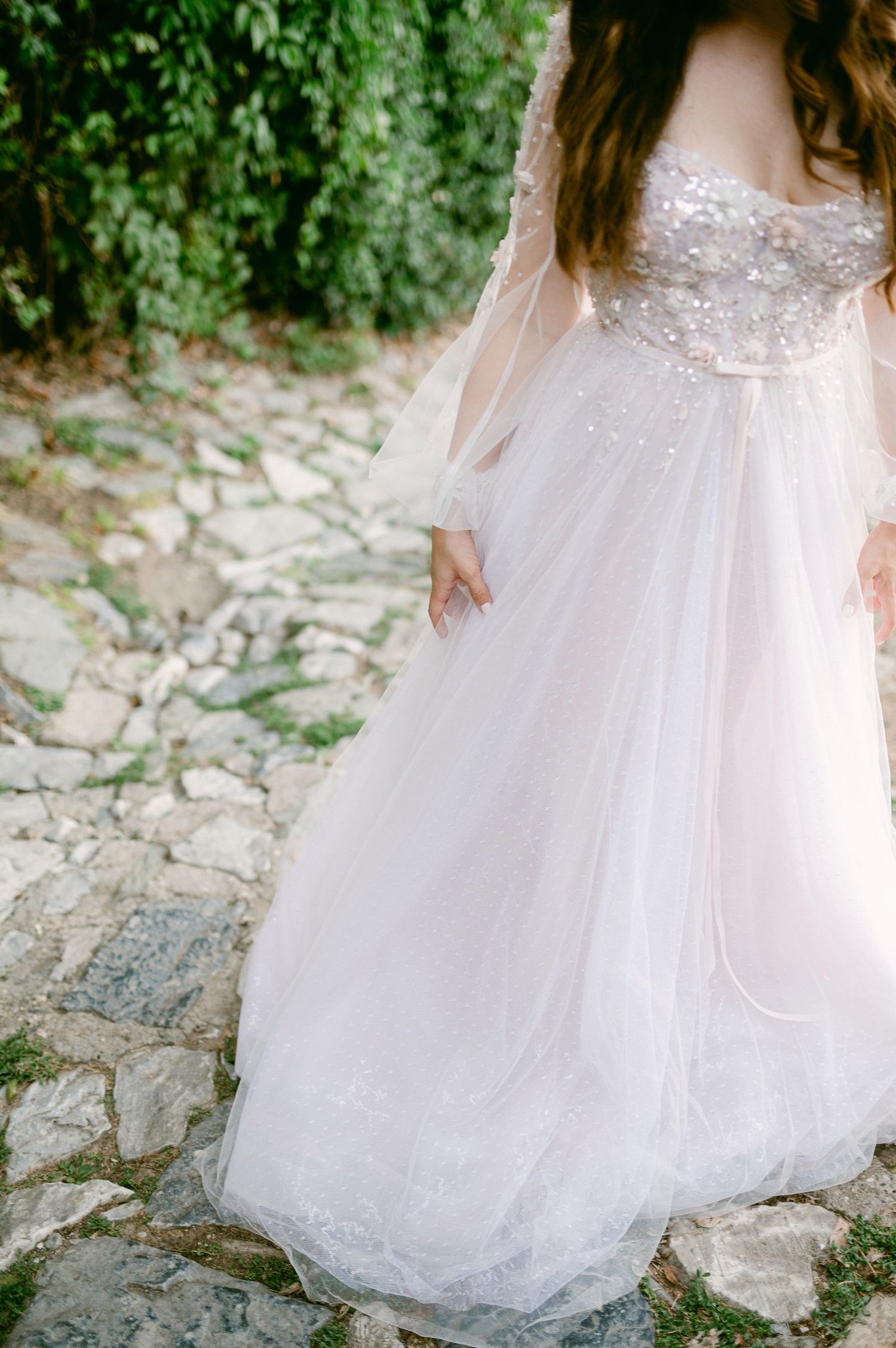 A flowing white wedding dress with lace details and sheer sleeves is photographed on a rustic stone path surrounded by greenery.