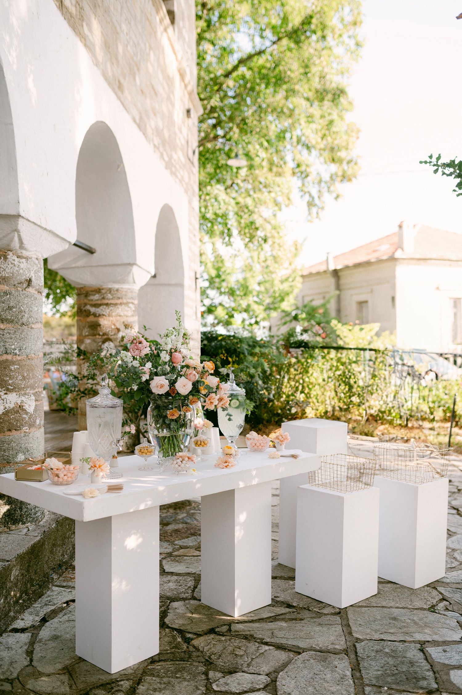 Elegant outdoor dining setup with white modern furniture and pink floral arrangements under Mediterranean-style arches.
