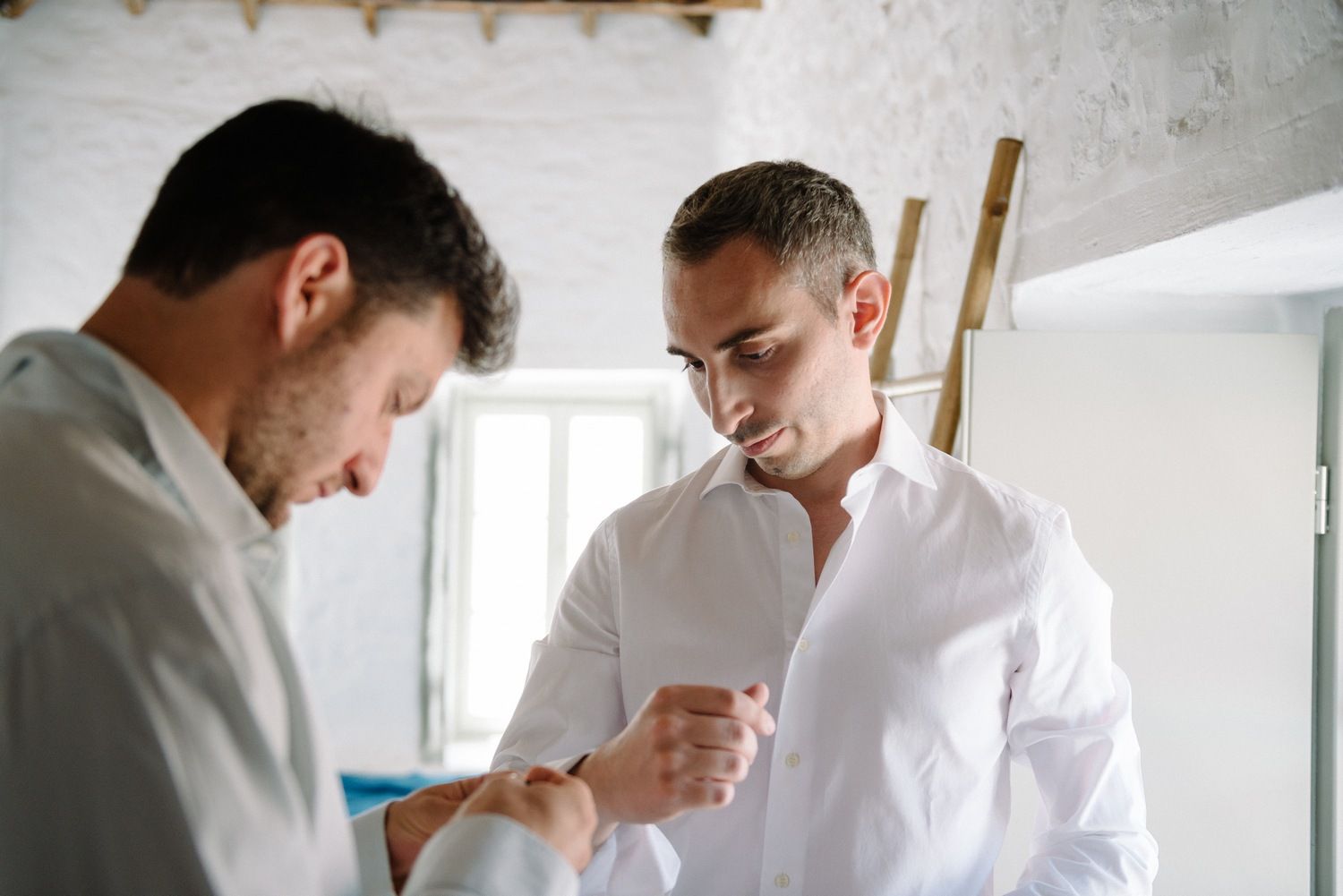 Two men in white shirts preparing formal attire in a bright interior space.