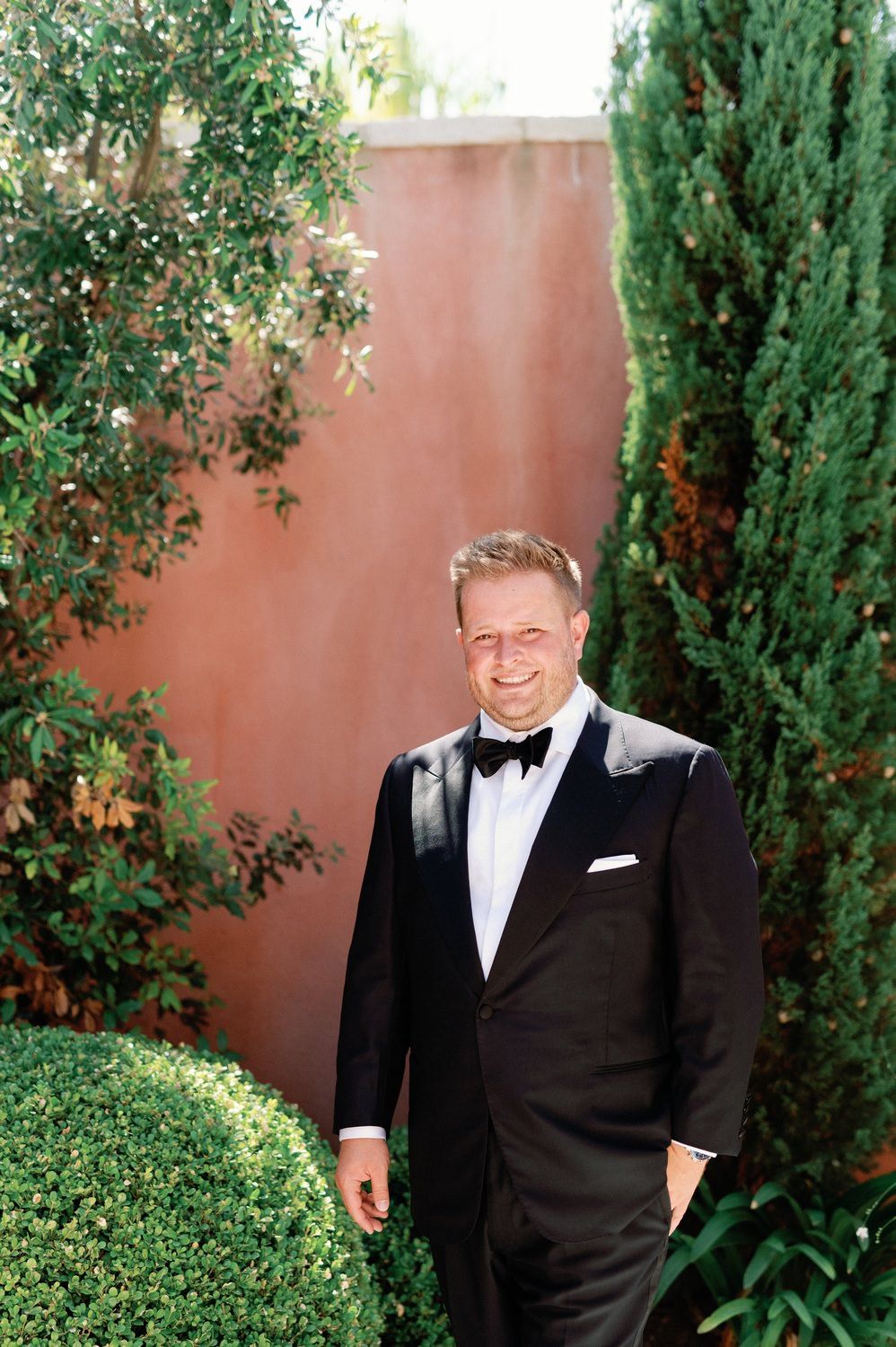 A person in a classic black tuxedo and bow tie smiles while standing in a garden with cypress trees and a terracotta wall.