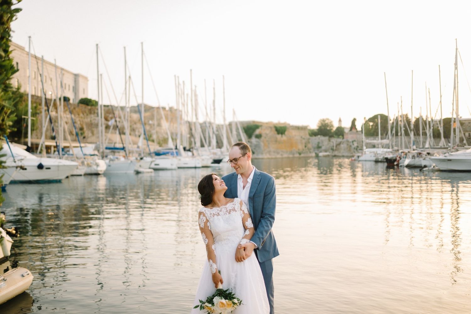 Romantic sunset portrait of newlyweds by marina with sailboats in background.