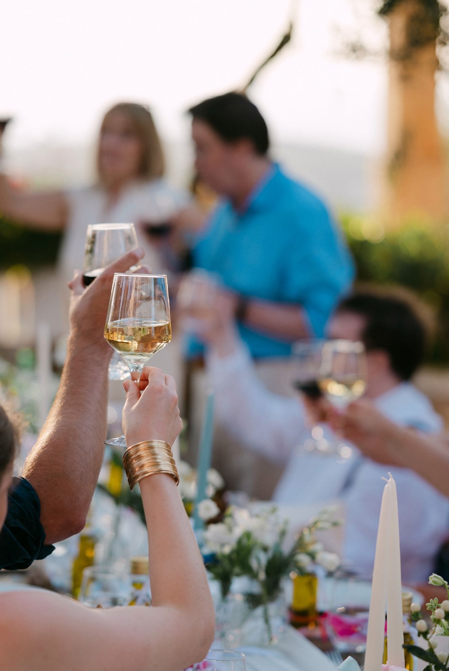 People raise wine glasses in a toast at an outdoor dinner party with floral centerpieces and candlelight.