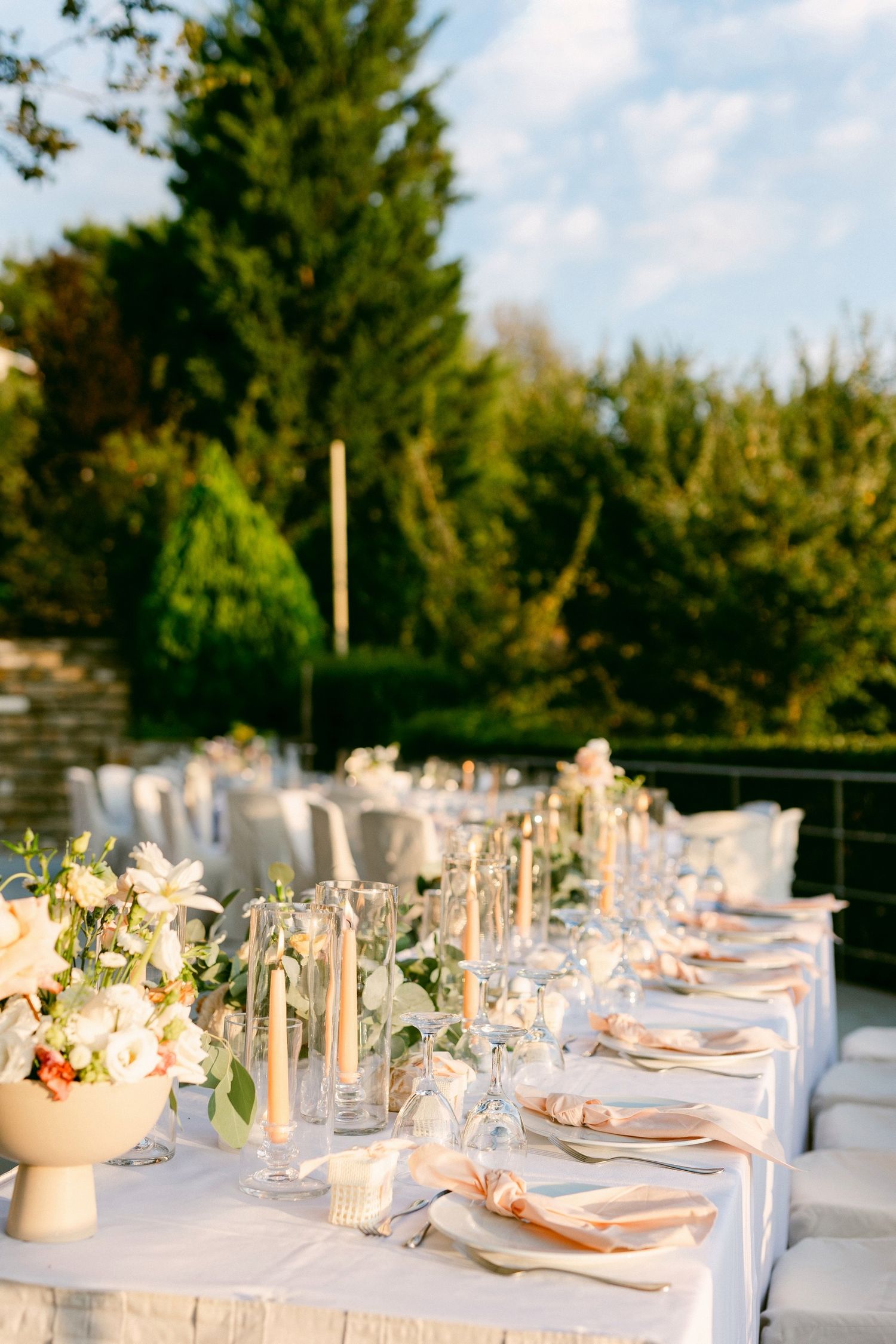 Elegant outdoor wedding reception table set with white linens, crystal glassware and delicate floral centerpieces at sunset.