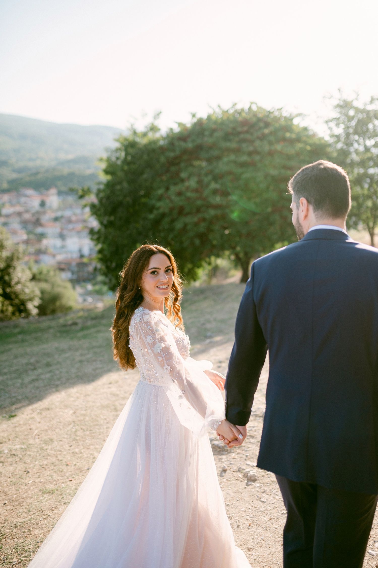 A couple in wedding attire walk hand in hand on a hilltop path with a scenic village view in the background.