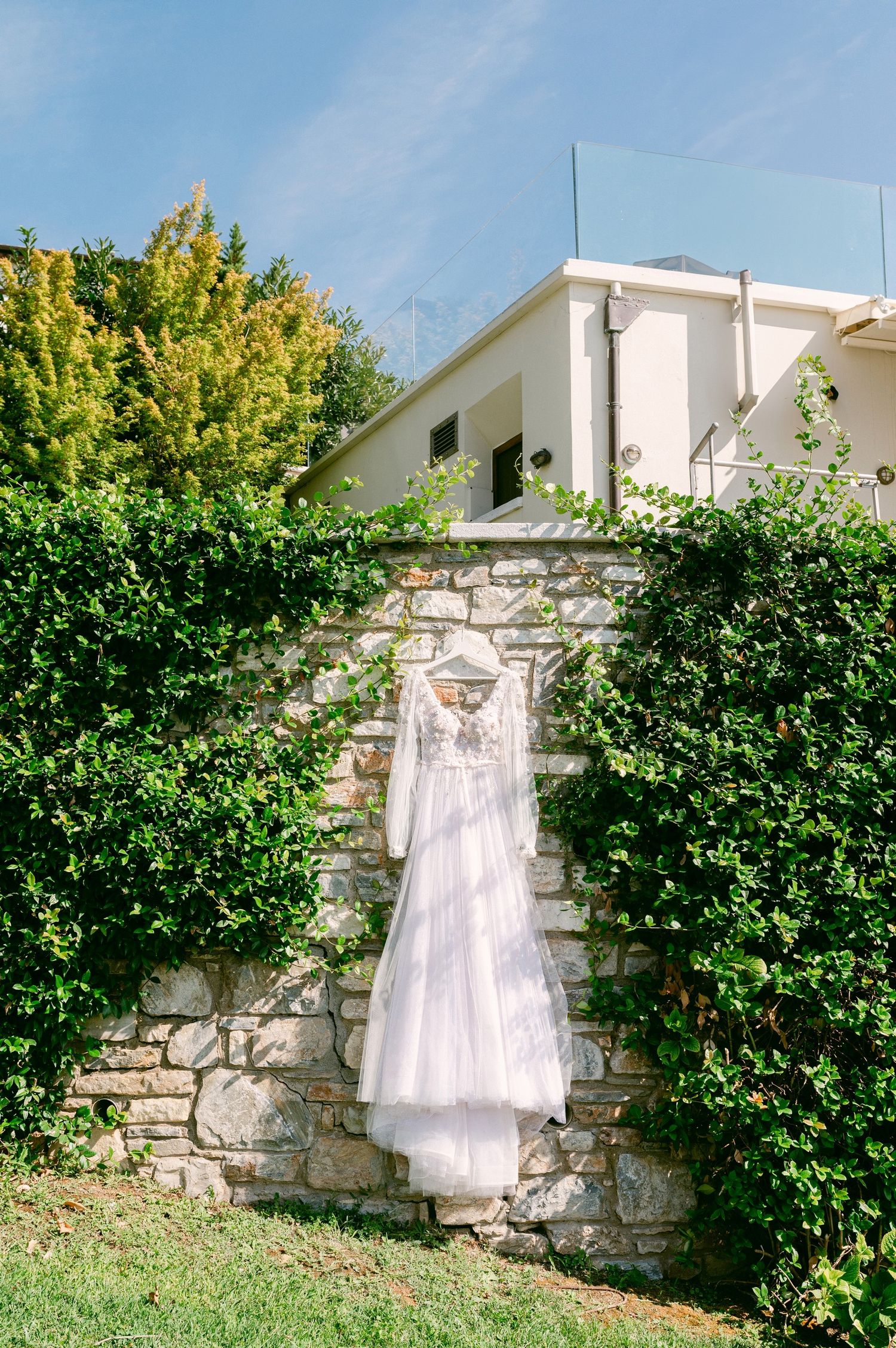 A white wedding dress hangs on a stone wall covered in green vines and foliage outside a Mediterranean-style building.