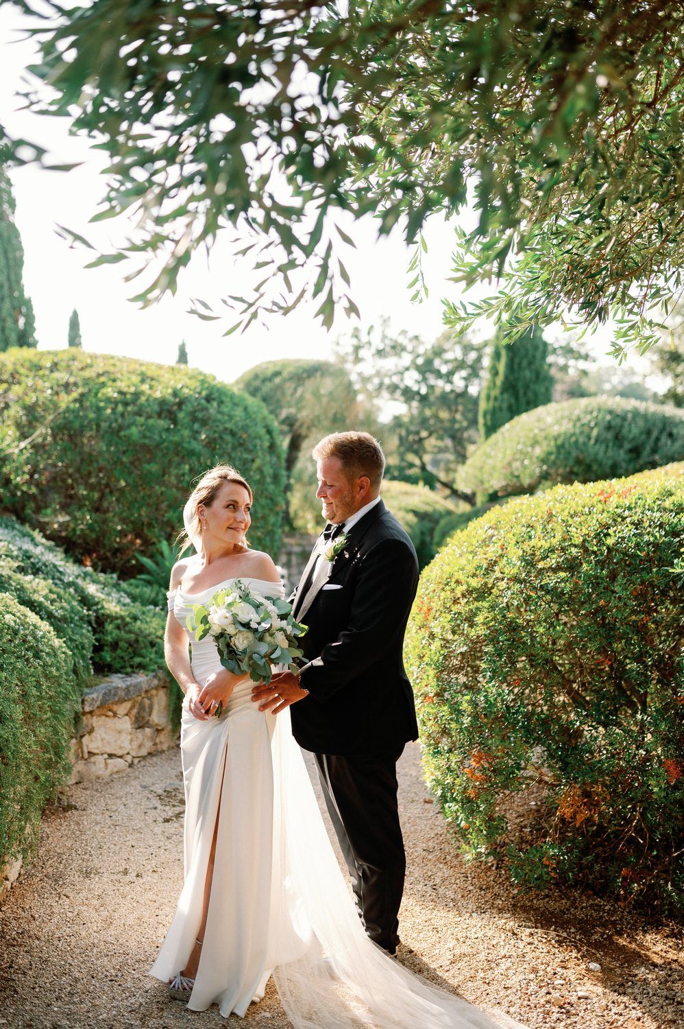 Newlyweds share intimate moment on garden path surrounded by manicured hedges.