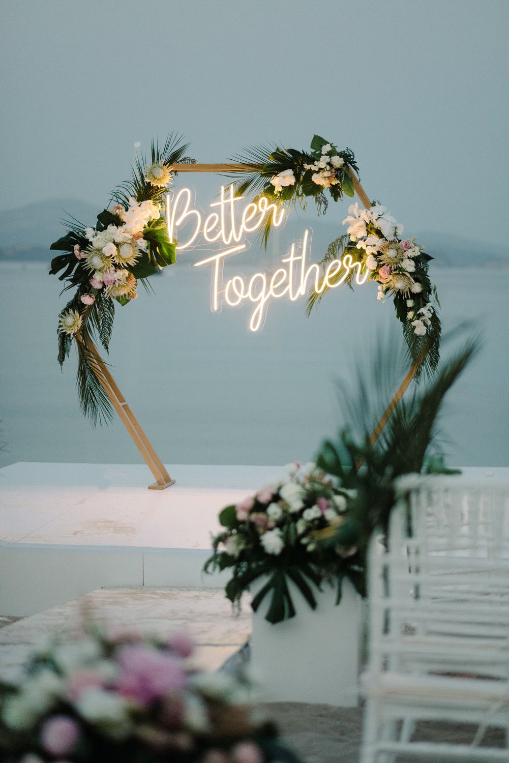Illuminated neon 'Better Together' sign adorned with tropical flowers and greenery against mountain backdrop.