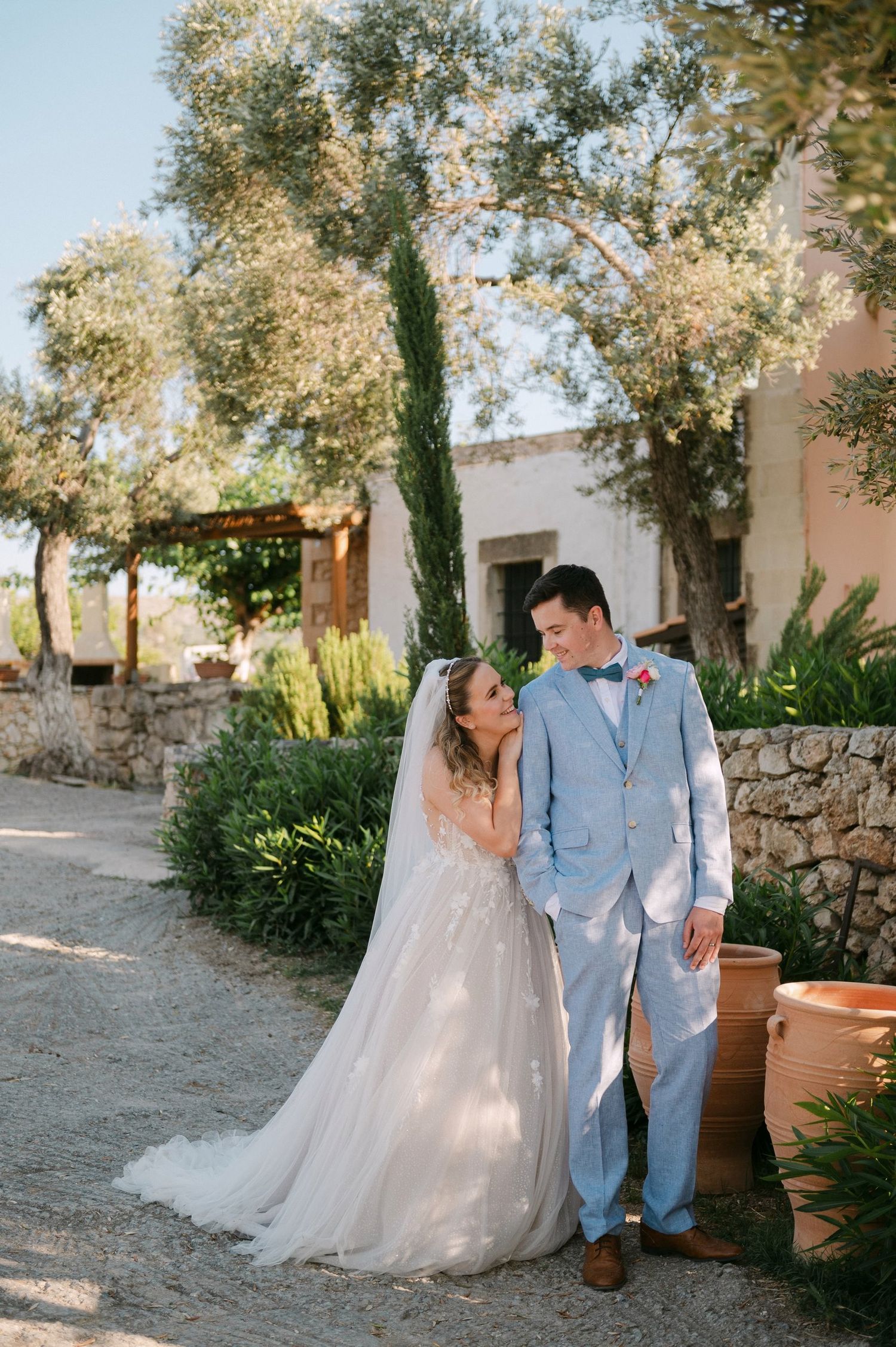 A couple shares a romantic moment in their wedding attire against a rustic stone wall with cypress trees and gardens.