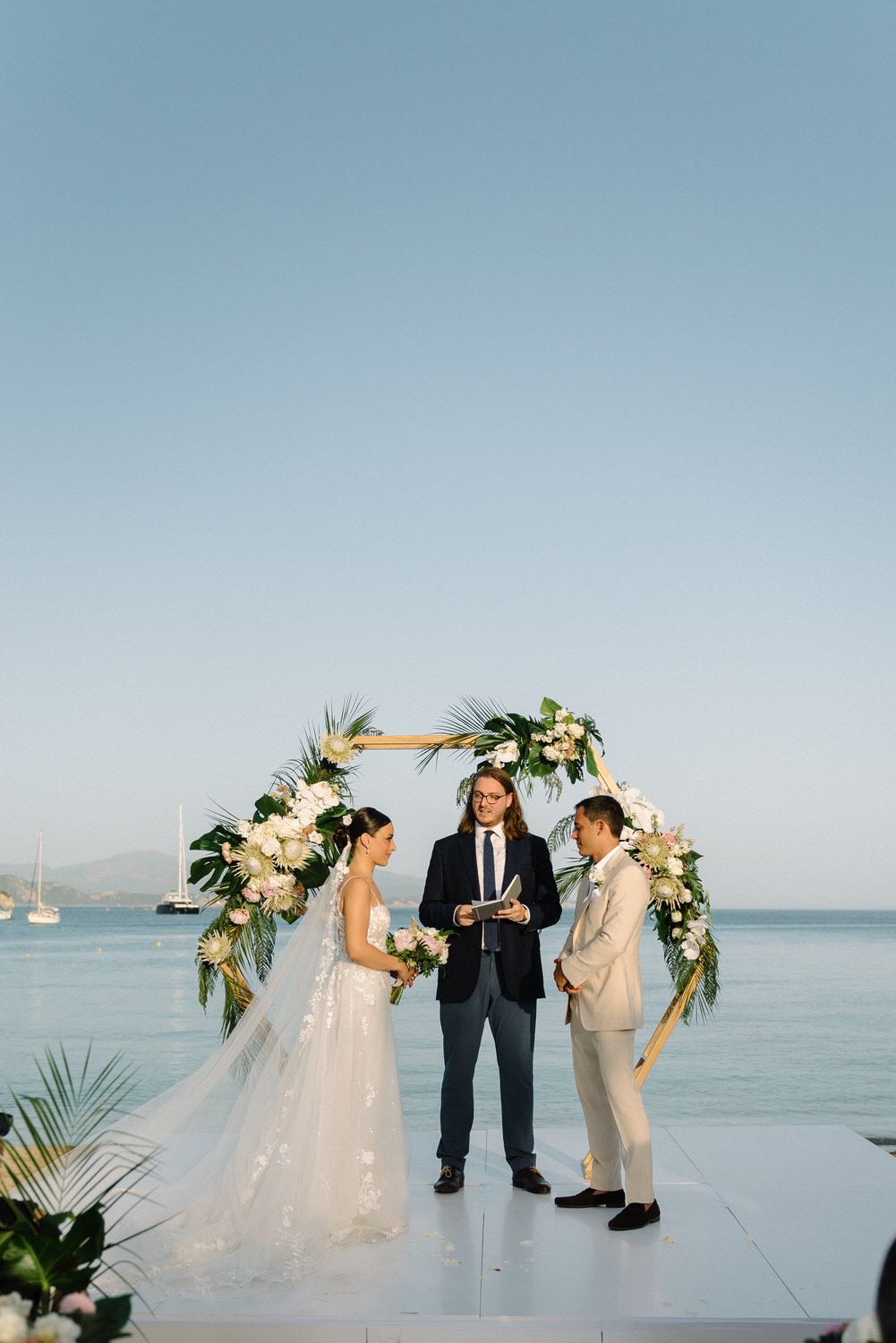 Elegant waterfront wedding ceremony with floral arch and mountains in the background.