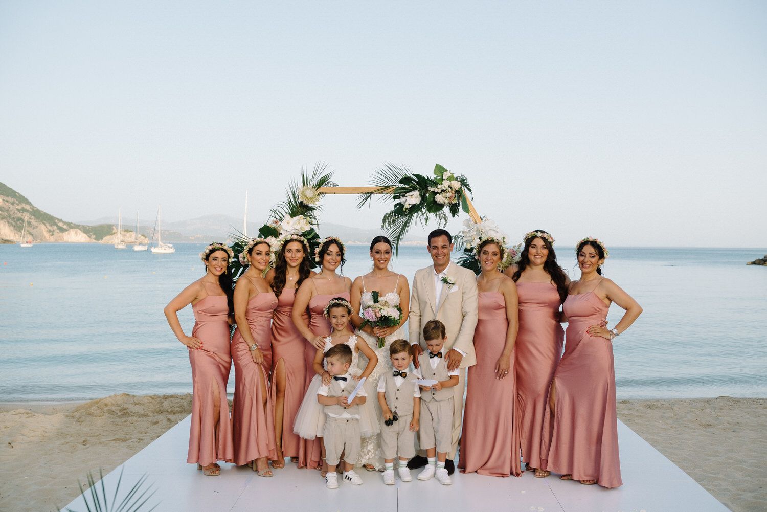 Wedding party poses together on beach platform wearing pink dresses and light-colored suits.