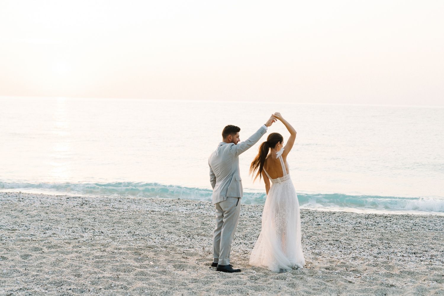 A couple in wedding attire dances on a beach at sunset with ocean waves in the background.