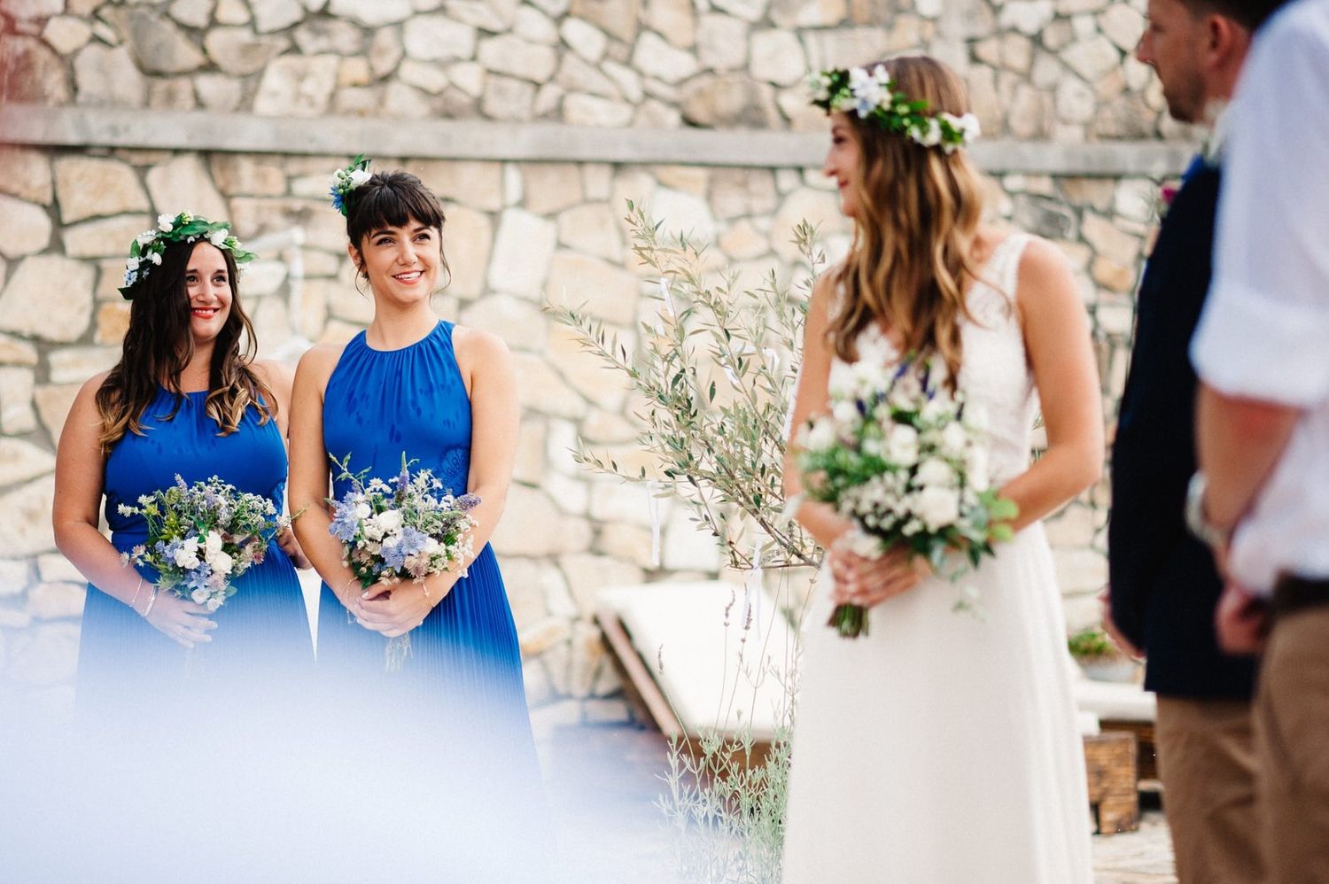 Bridesmaids in vibrant blue dresses hold bouquets while standing near a stone wall at an outdoor wedding ceremony.