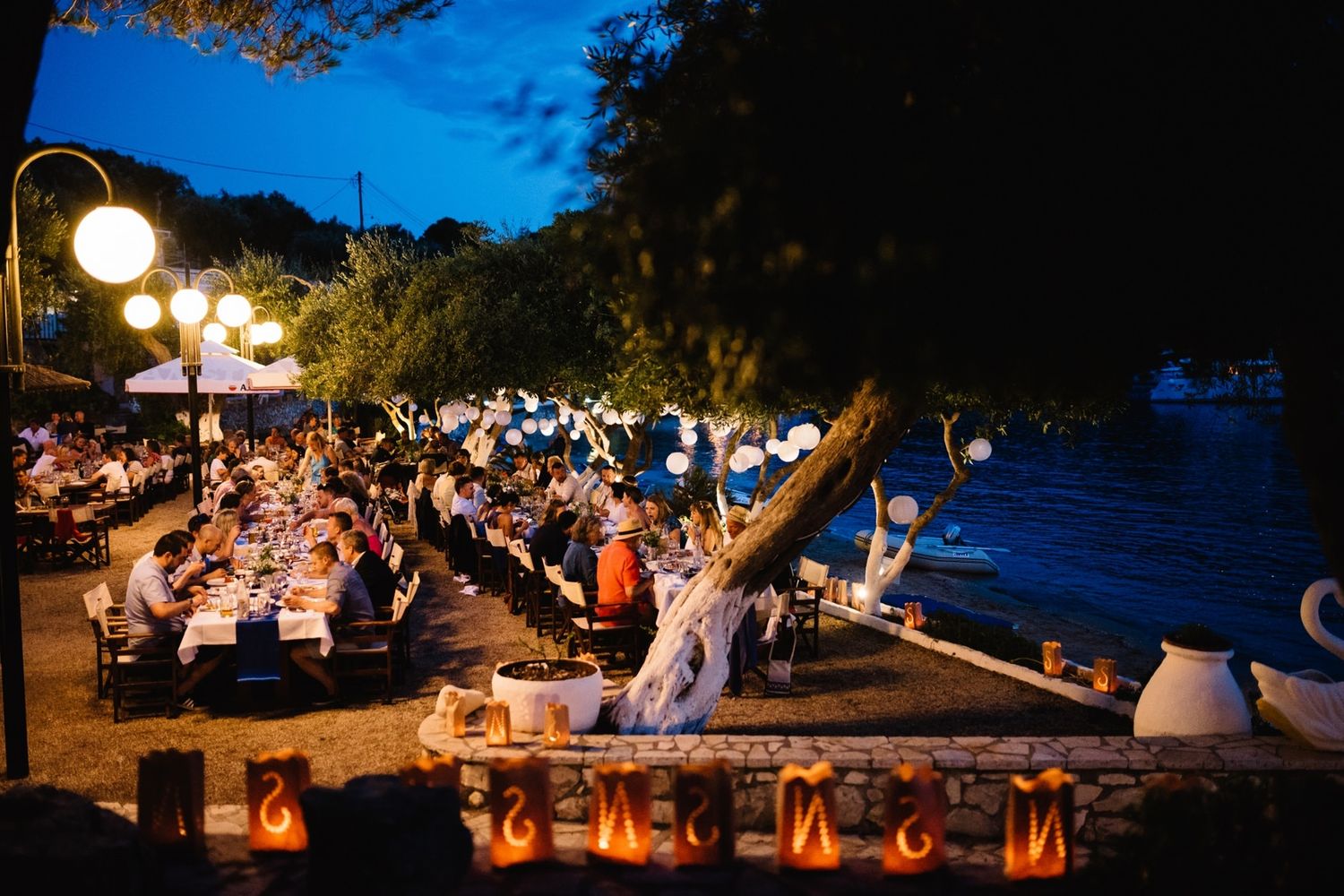 Evening outdoor wedding reception with long tables and string lights by the water.