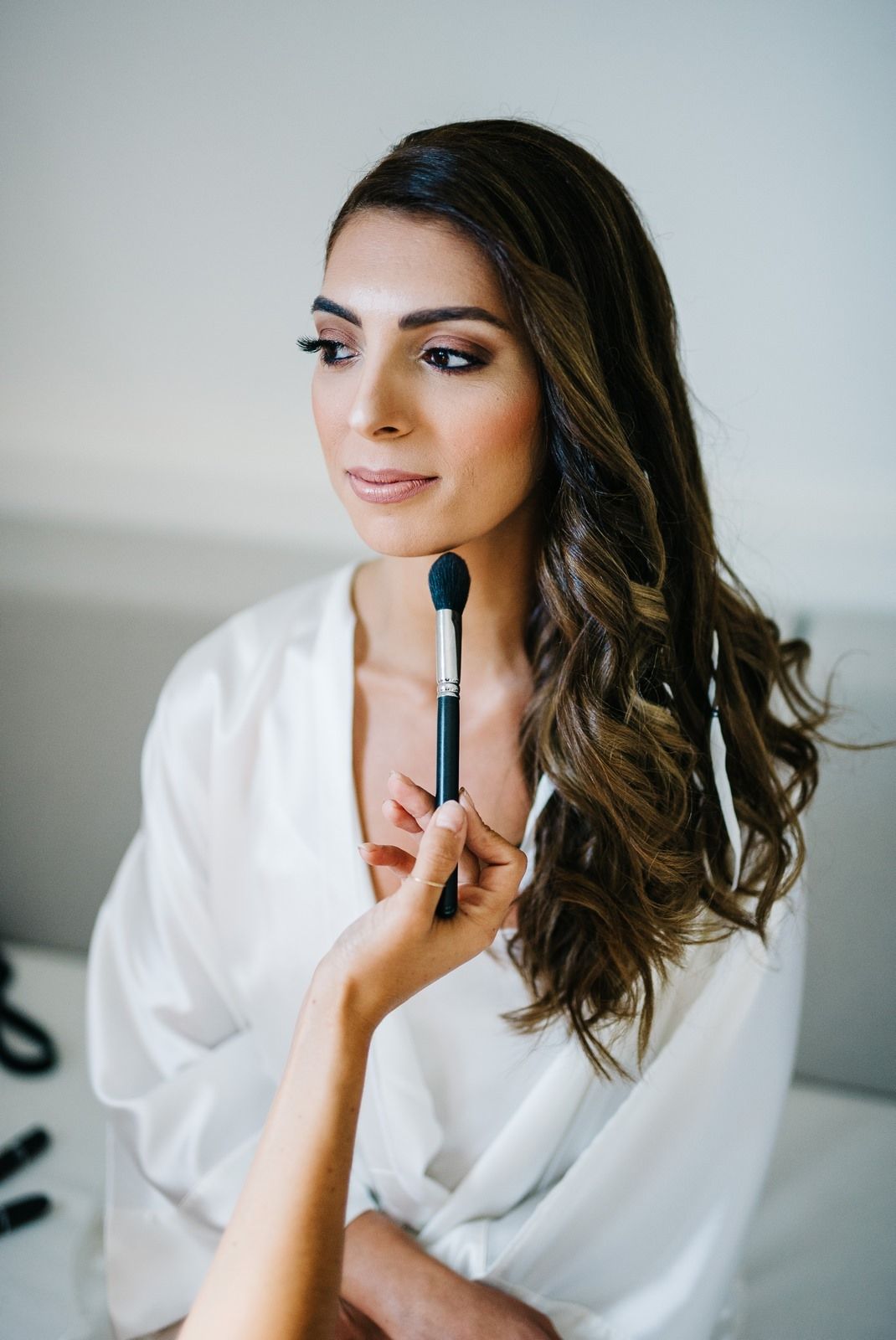 Professional makeup application in progress with white silk robe in natural lighting.