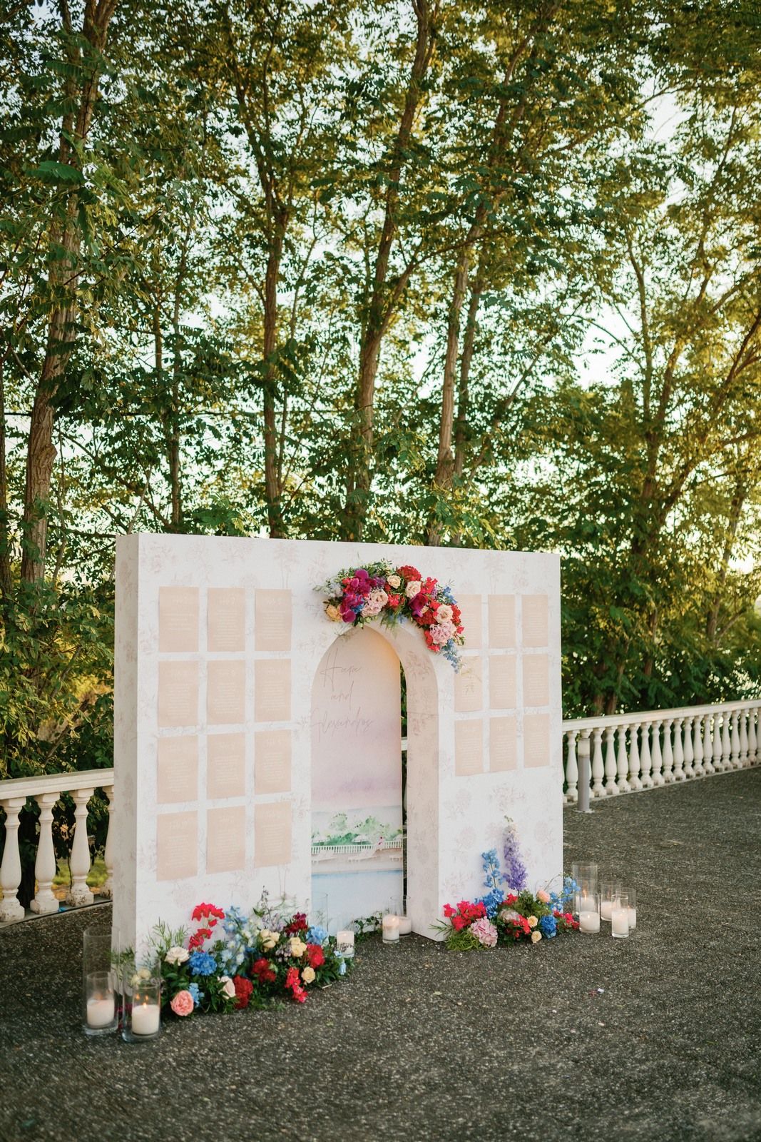 Elegant white wedding arch decorated with pink and red flowers against a backdrop of tall trees at sunset.