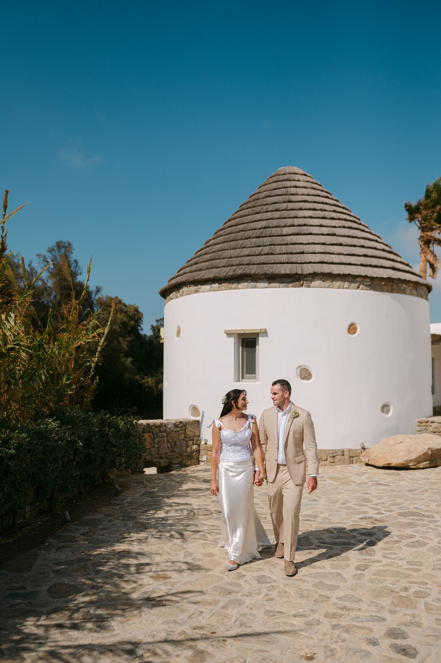 A couple walks together on a stone path next to a white circular building with a conical roof in Mediterranean setting.