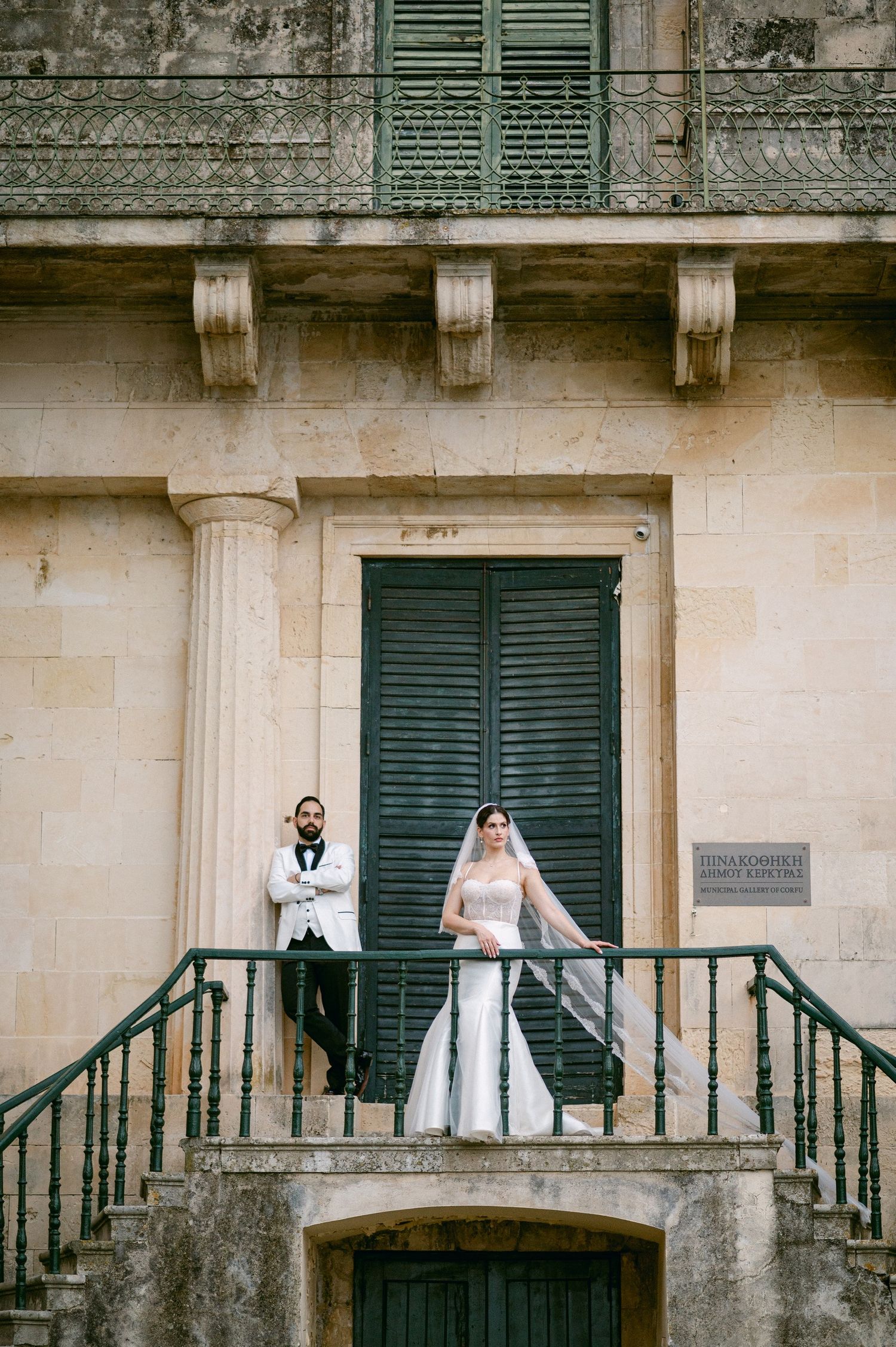 A historic building balcony with green shutters and stone columns shows two people in white attire standing outside.