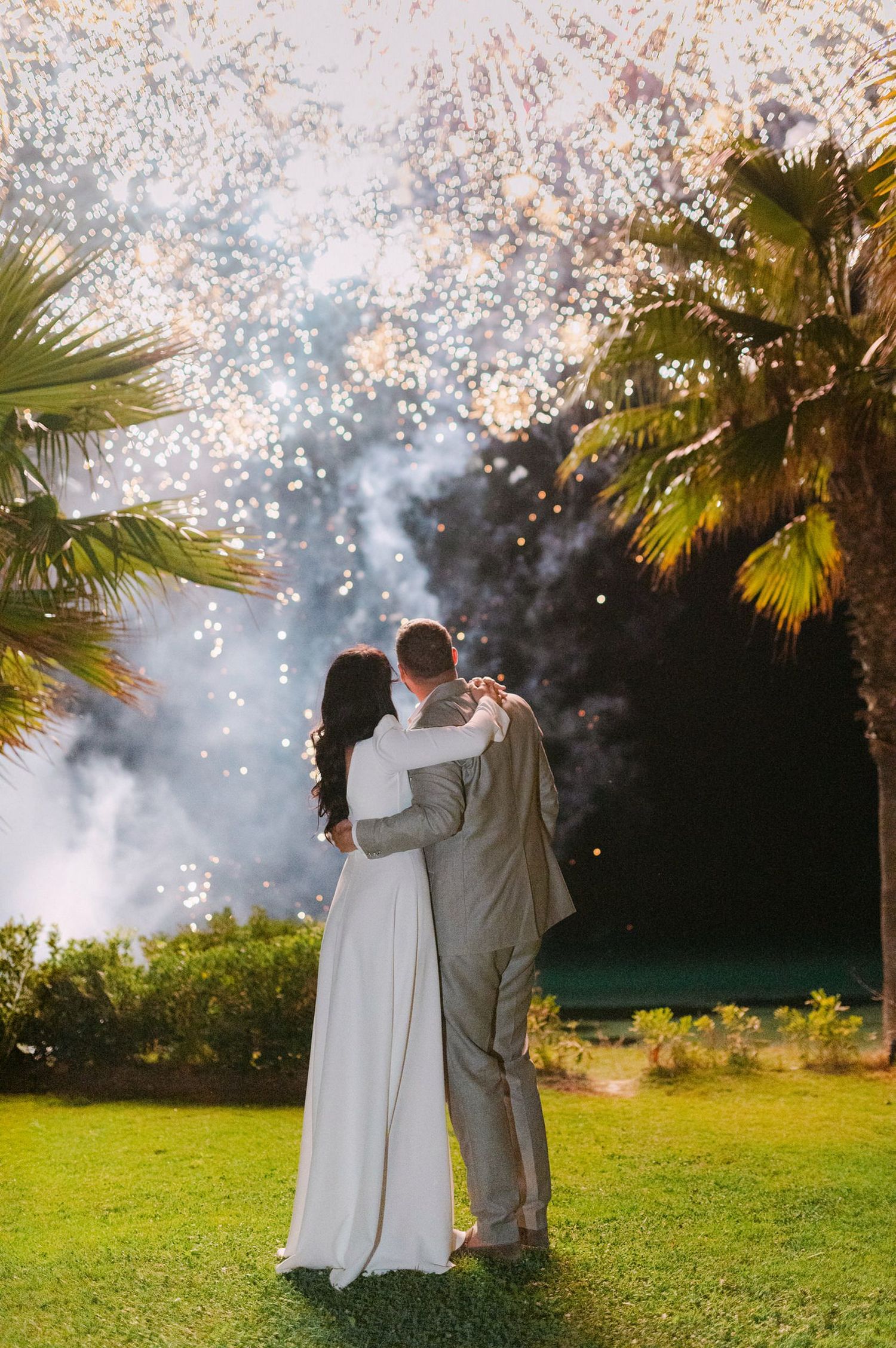A couple embraces on a lawn while fireworks sparkle in the evening sky behind palm trees.