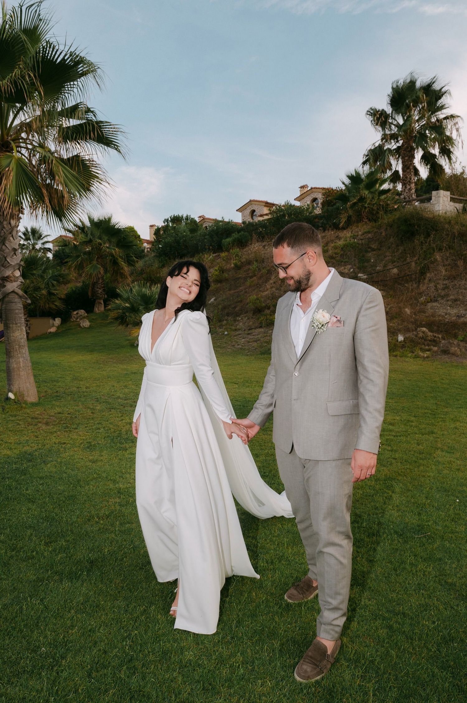 A couple in wedding attire holds hands on a grassy hillside with palm trees and houses in the background.