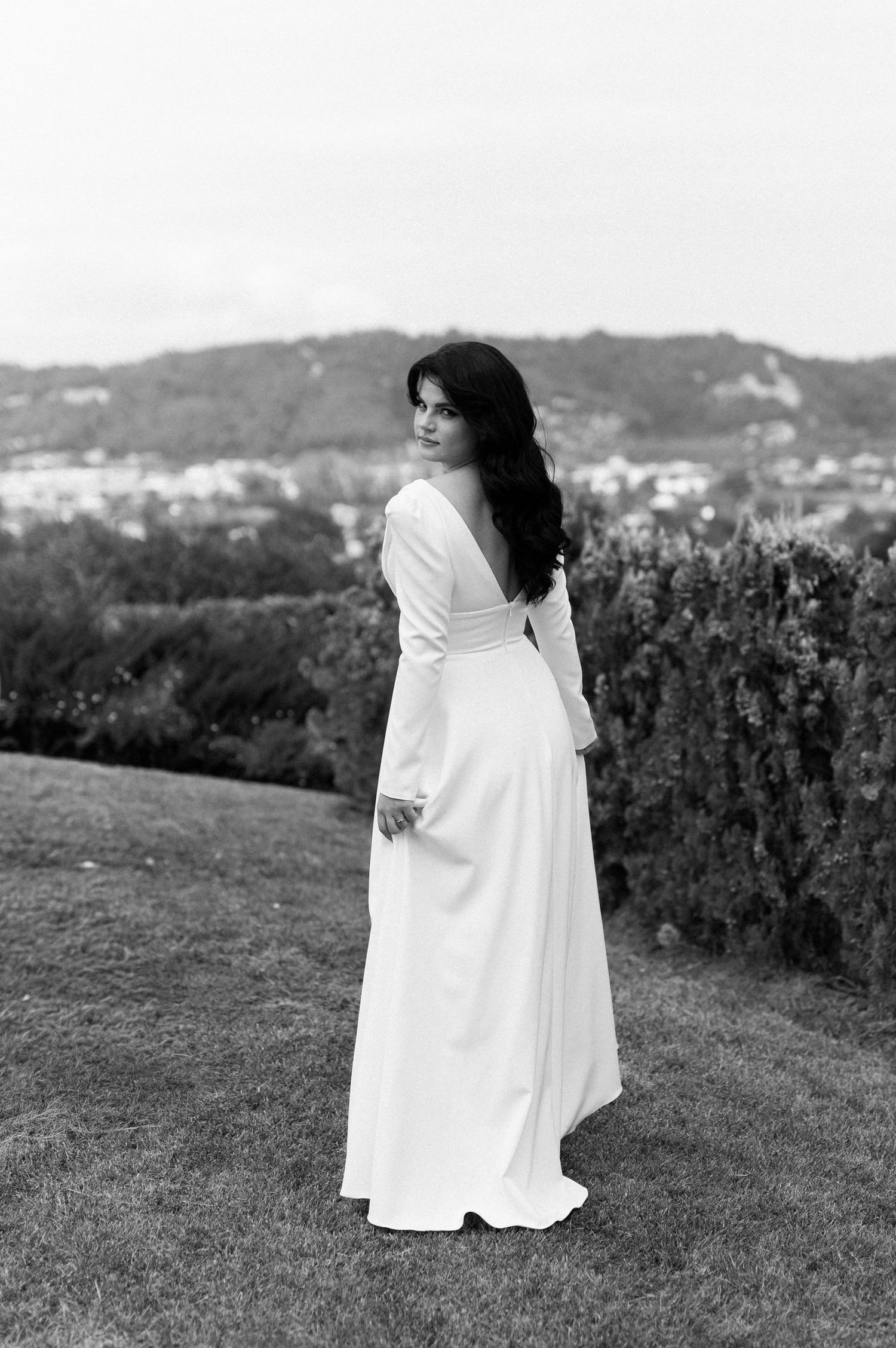 A person in a flowing white dress stands gracefully on a grassy hilltop with mountains in the background.