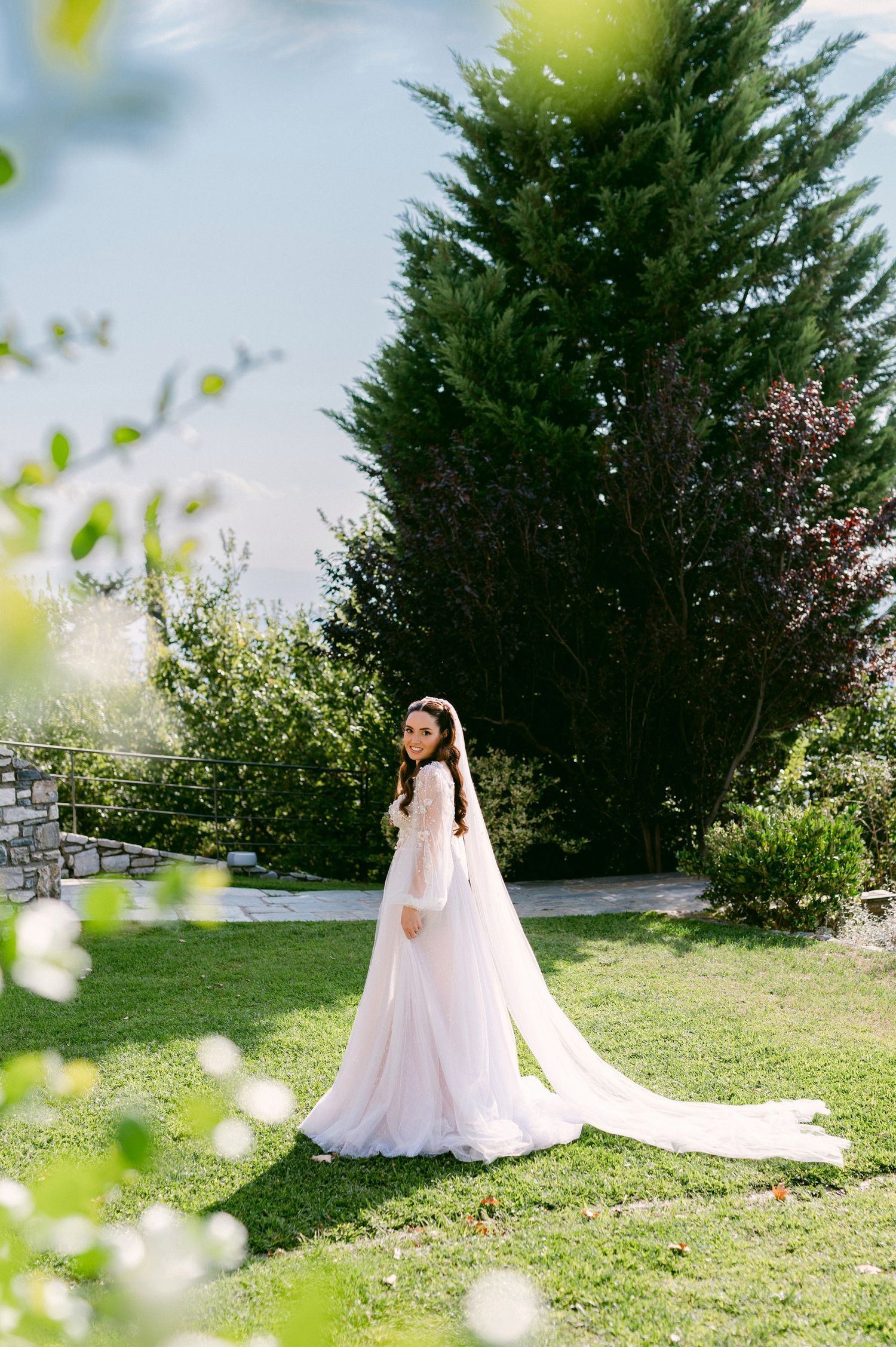 A bride in a flowing white dress and train stands on a sunny garden lawn with evergreen trees in the background.