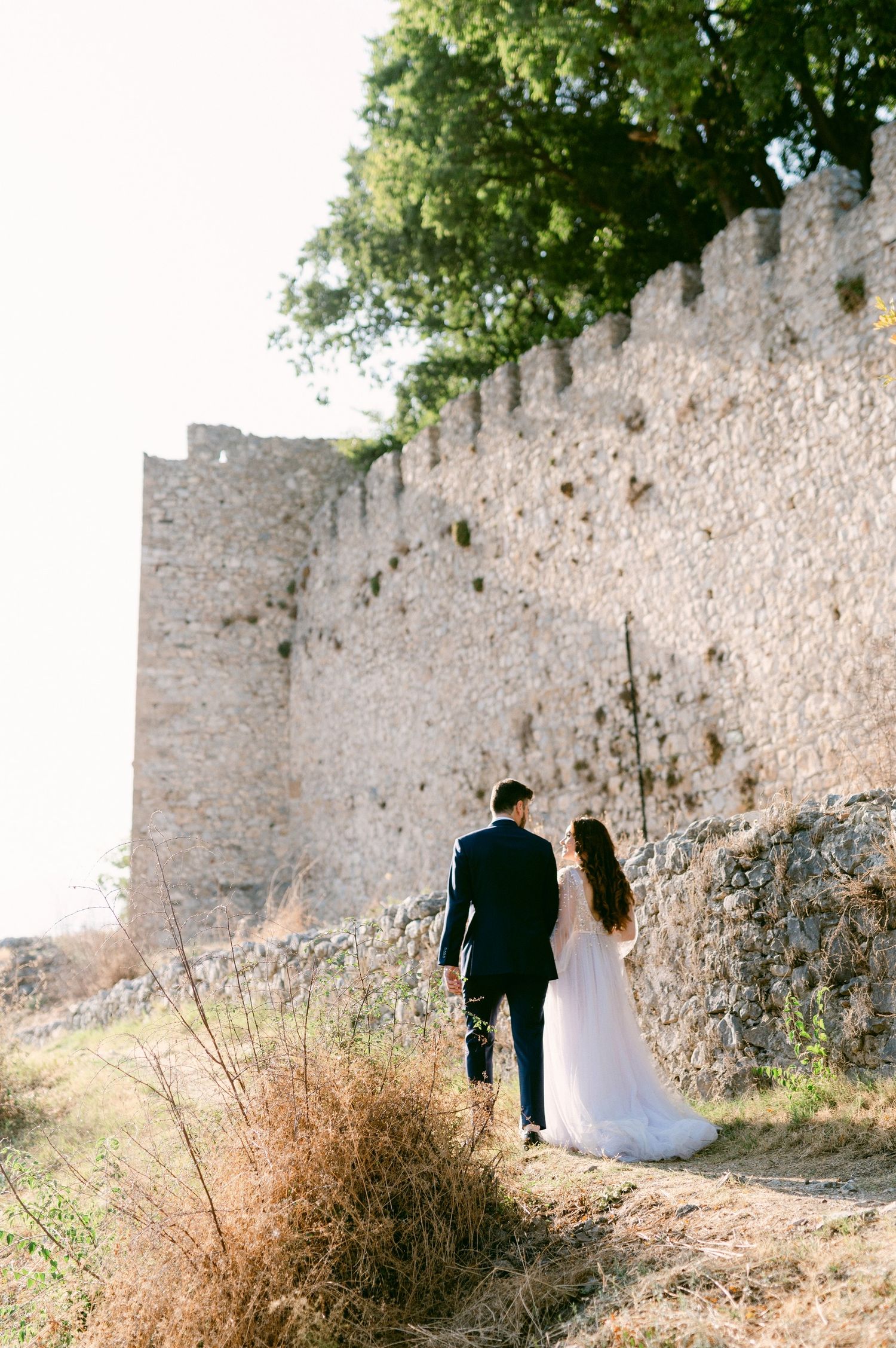 A couple in wedding attire walking along a path by an ancient stone castle wall on a sunny day.