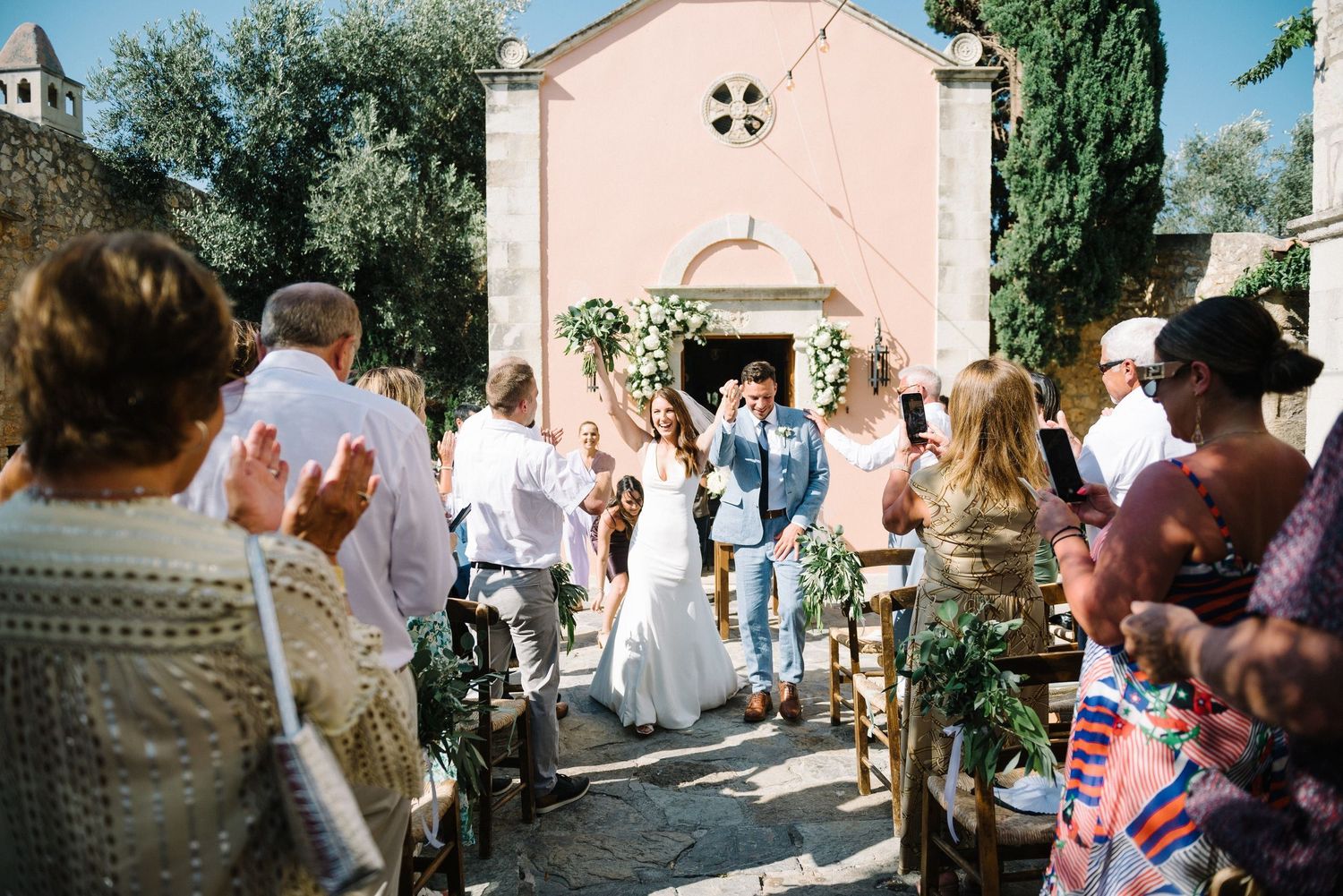 Wedding guests gather outside a charming pink church on a sunny summer day in Agreco Farms Crete.