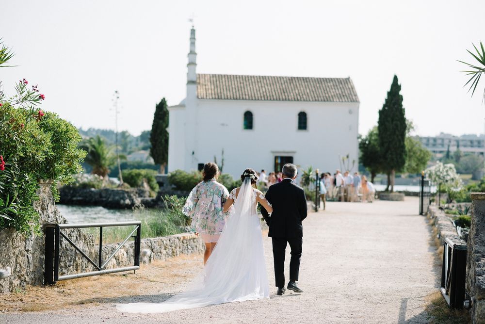 Wedding couple walks down a scenic path along a white Mediterranean church overlooking the sea in Corfu island.