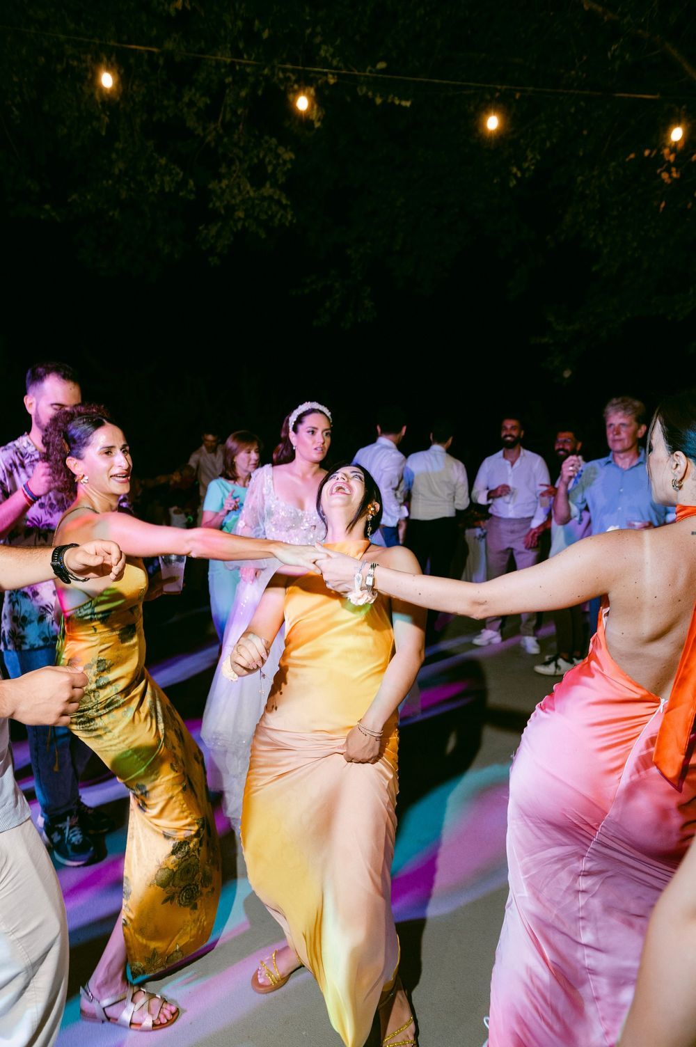 Wedding guests form a conga line dancing outdoors at night under string lights in colorful formal attire.