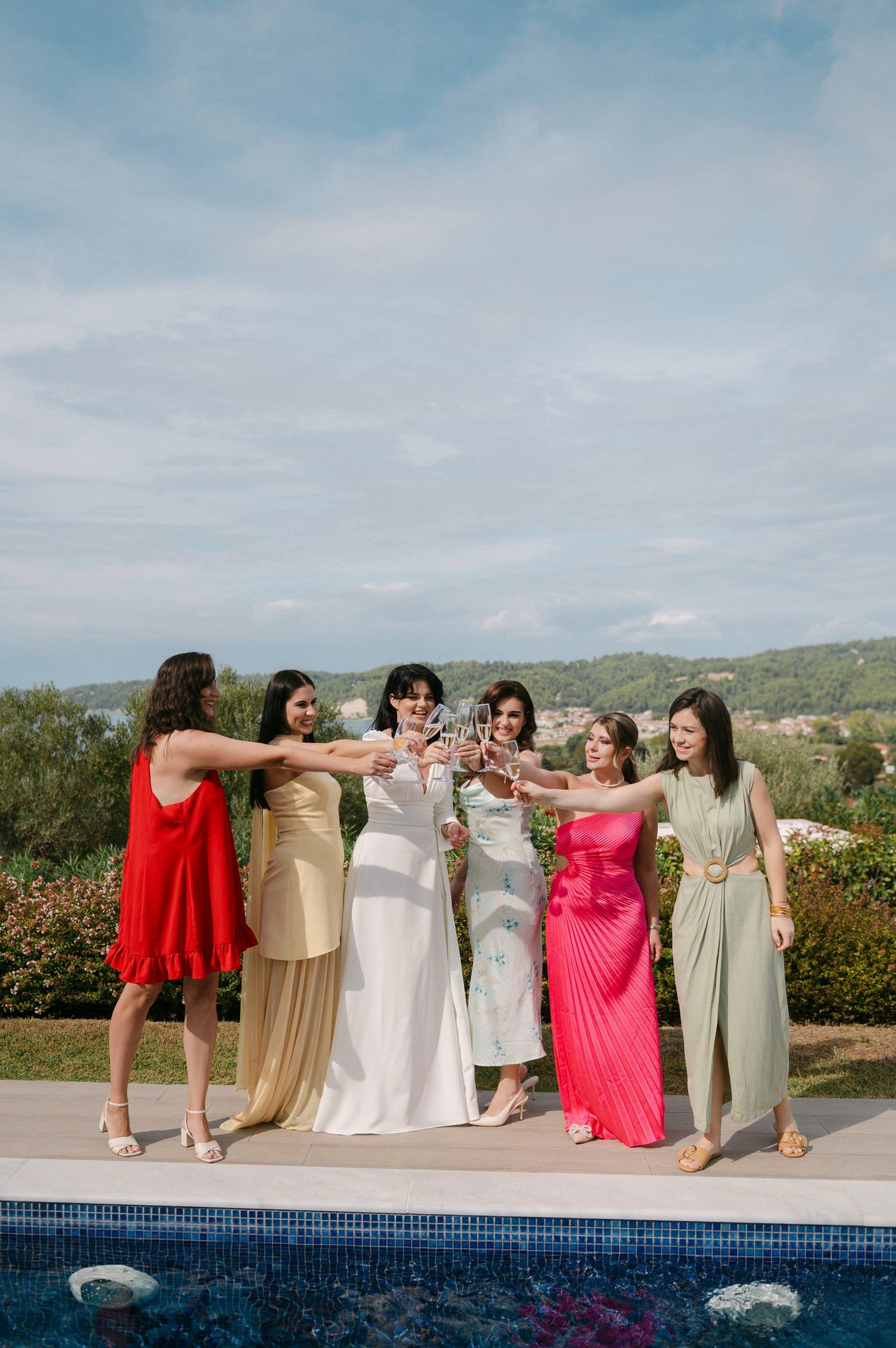 A group of women in coordinated wedding attire pose together by a swimming pool with scenic mountain views.