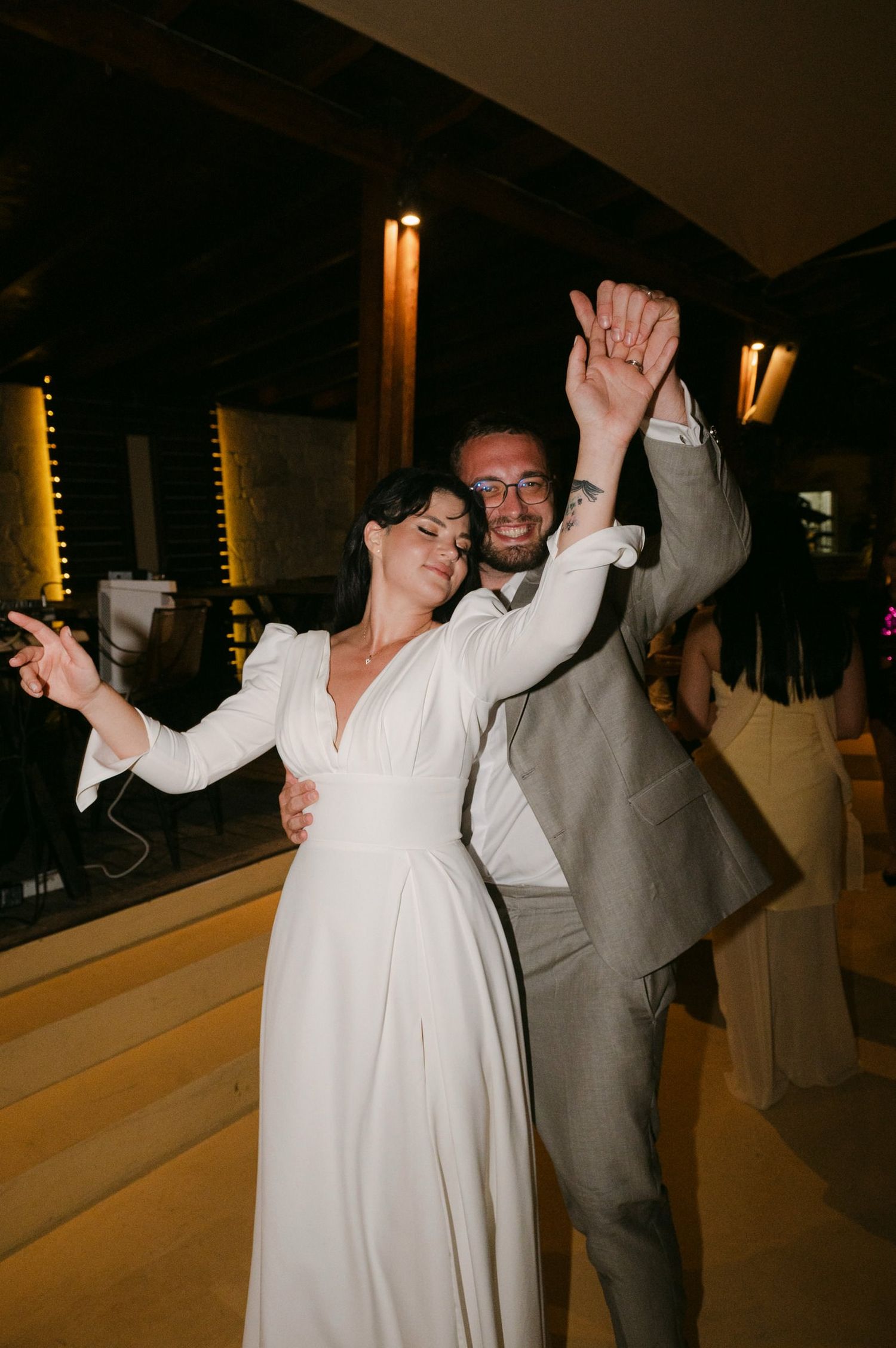 A couple dances together at a wedding reception, with the bride in white and groom in gray raising their arms joyfully.