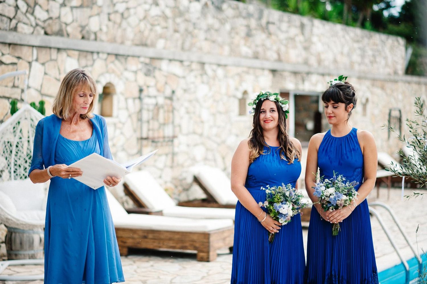 Wedding ceremony participants in blue dresses gather near a stone wall with lounge chairs.