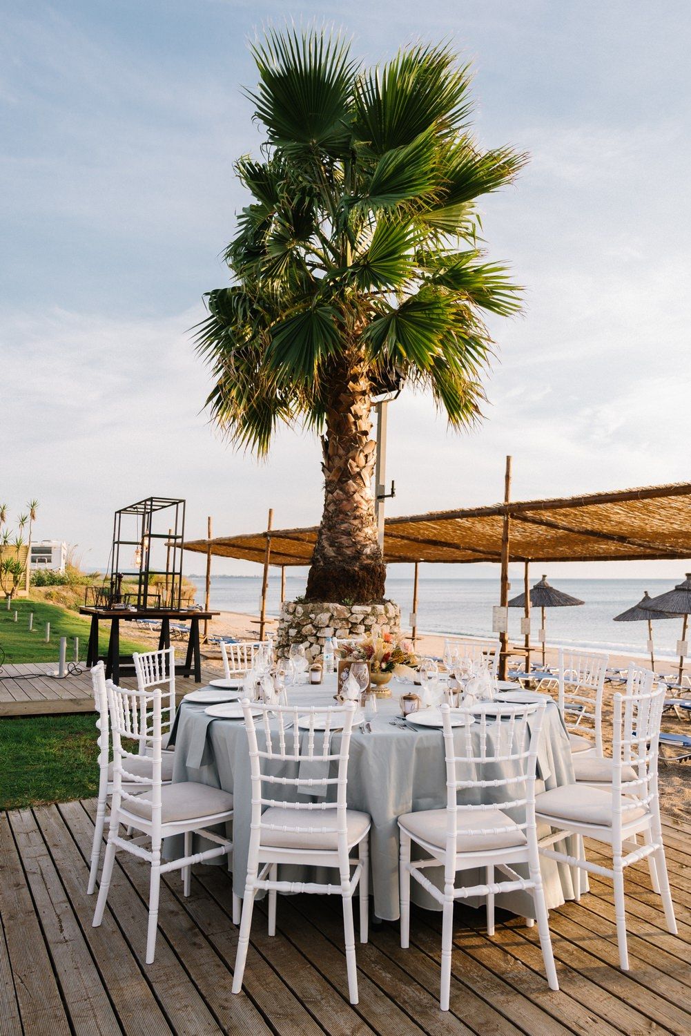 Outdoor wedding reception setup with white chairs around tables under a palm tree by the seaside.