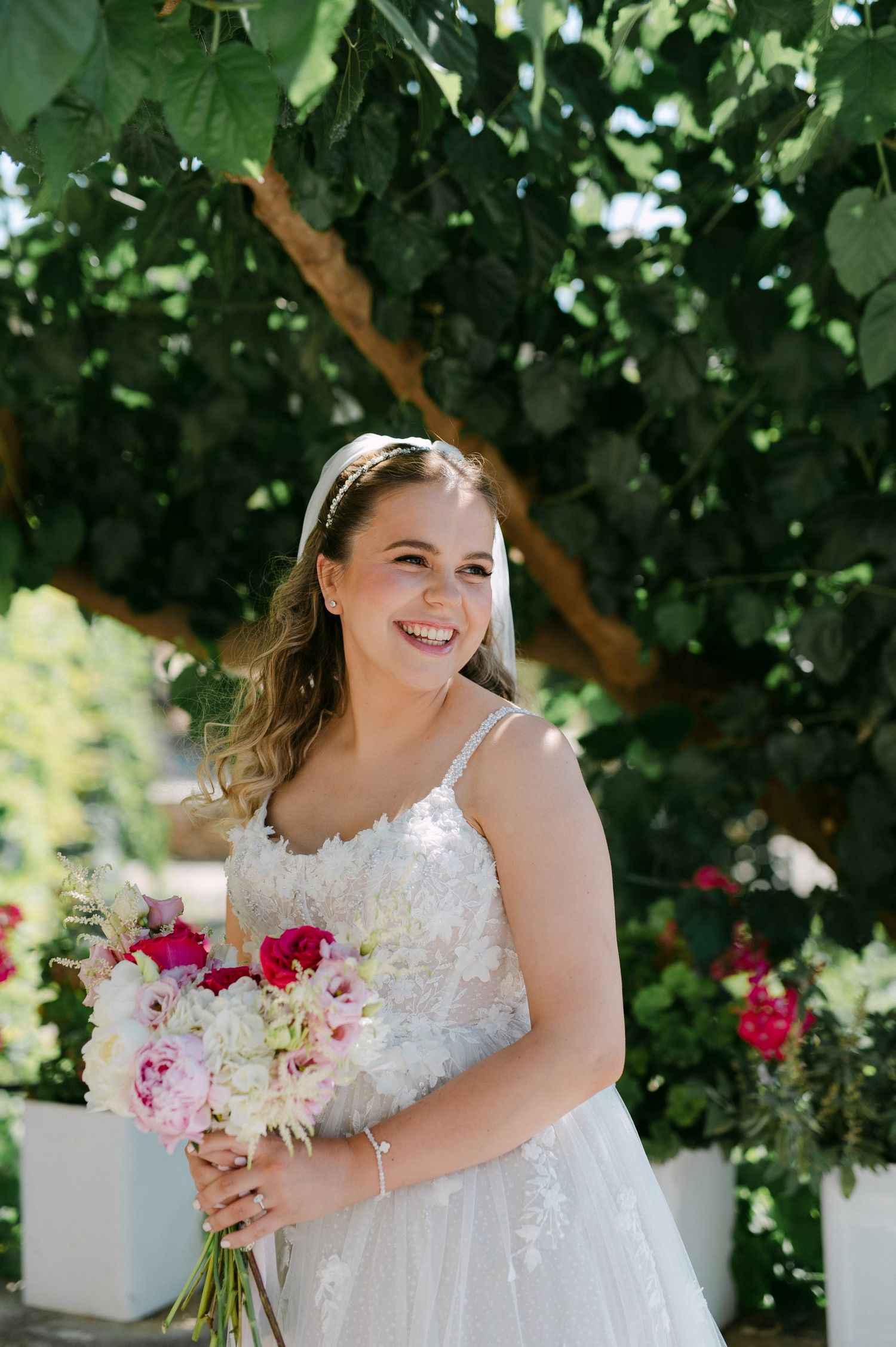 Bride in lace wedding dress poses with pink and white floral bouquet under garden trees on sunny day.