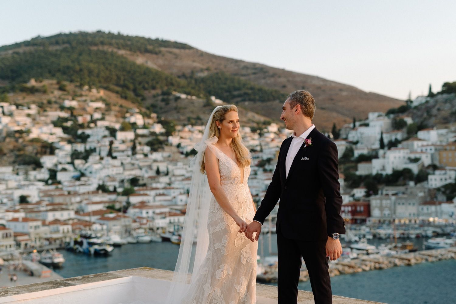 Couple in wedding attire holding hands on terrace with scenic Mediterranean village and boats in marina below.