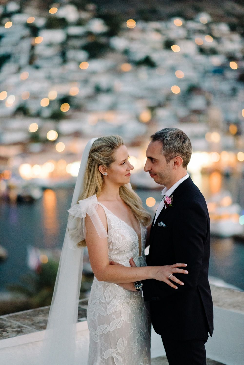 Newlyweds embrace on white terrace with coastal village lighting up behind them.