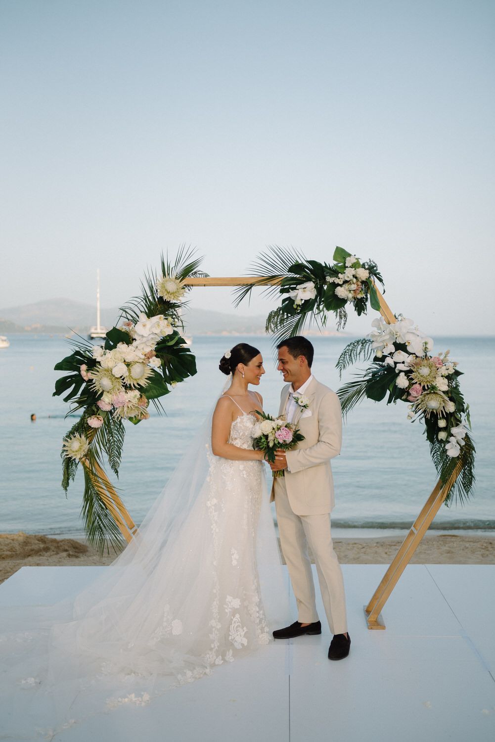 Romantic couple stands together under tropical floral arch at beach wedding ceremony.