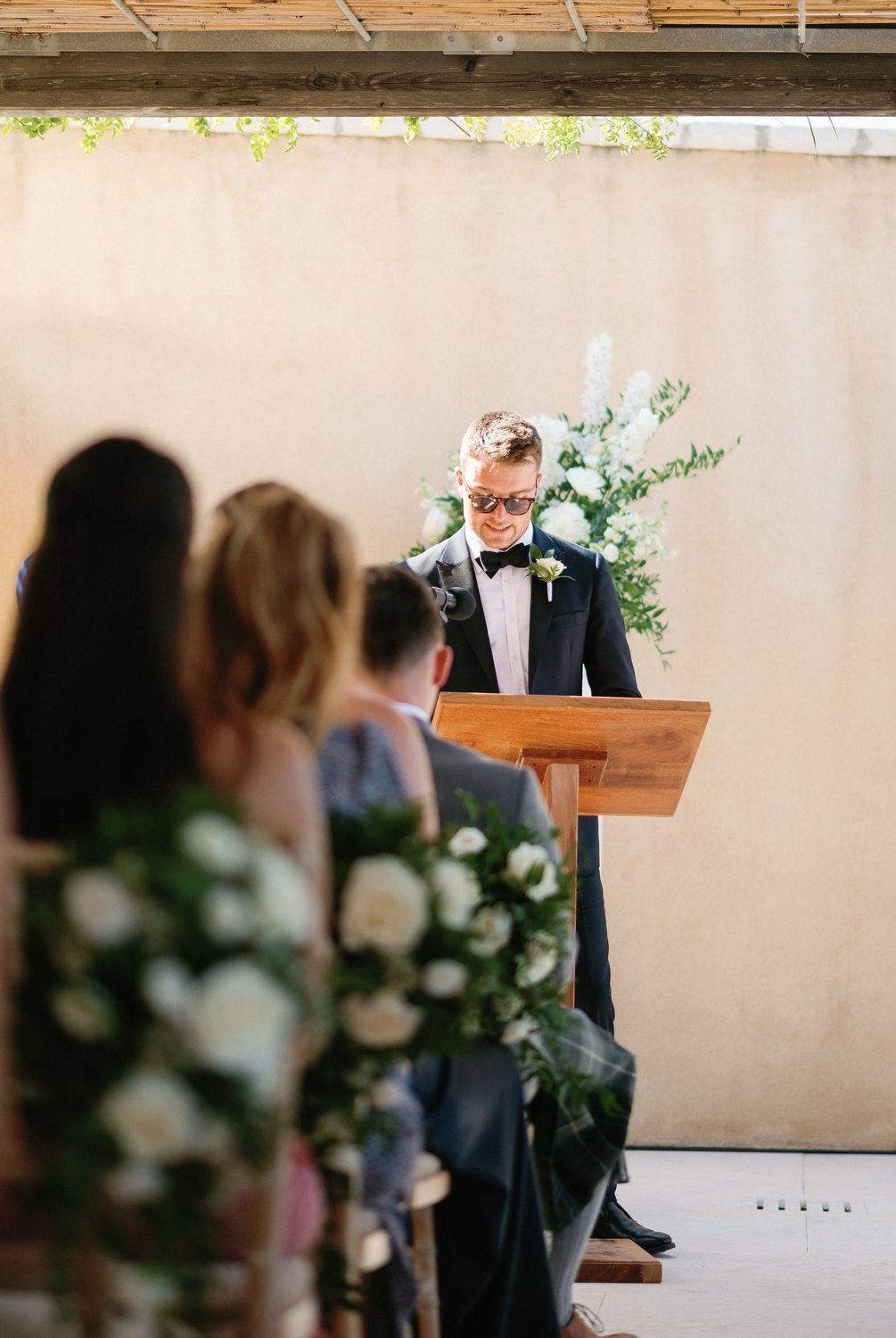 Wedding officiant speaking at wooden podium decorated with white floral arrangements.