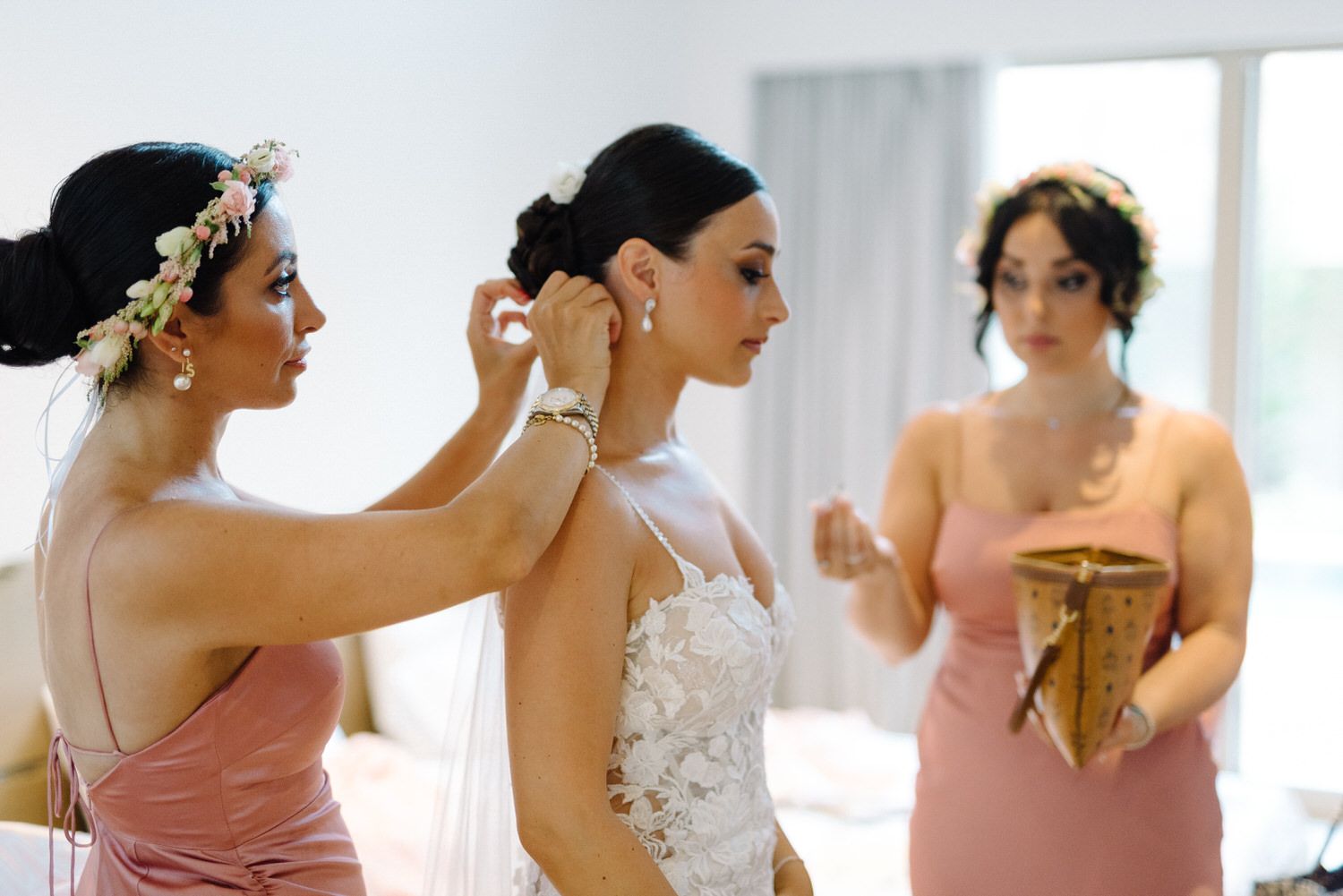 Bridesmaids in pink dresses helping with final wedding preparations in a bright room.