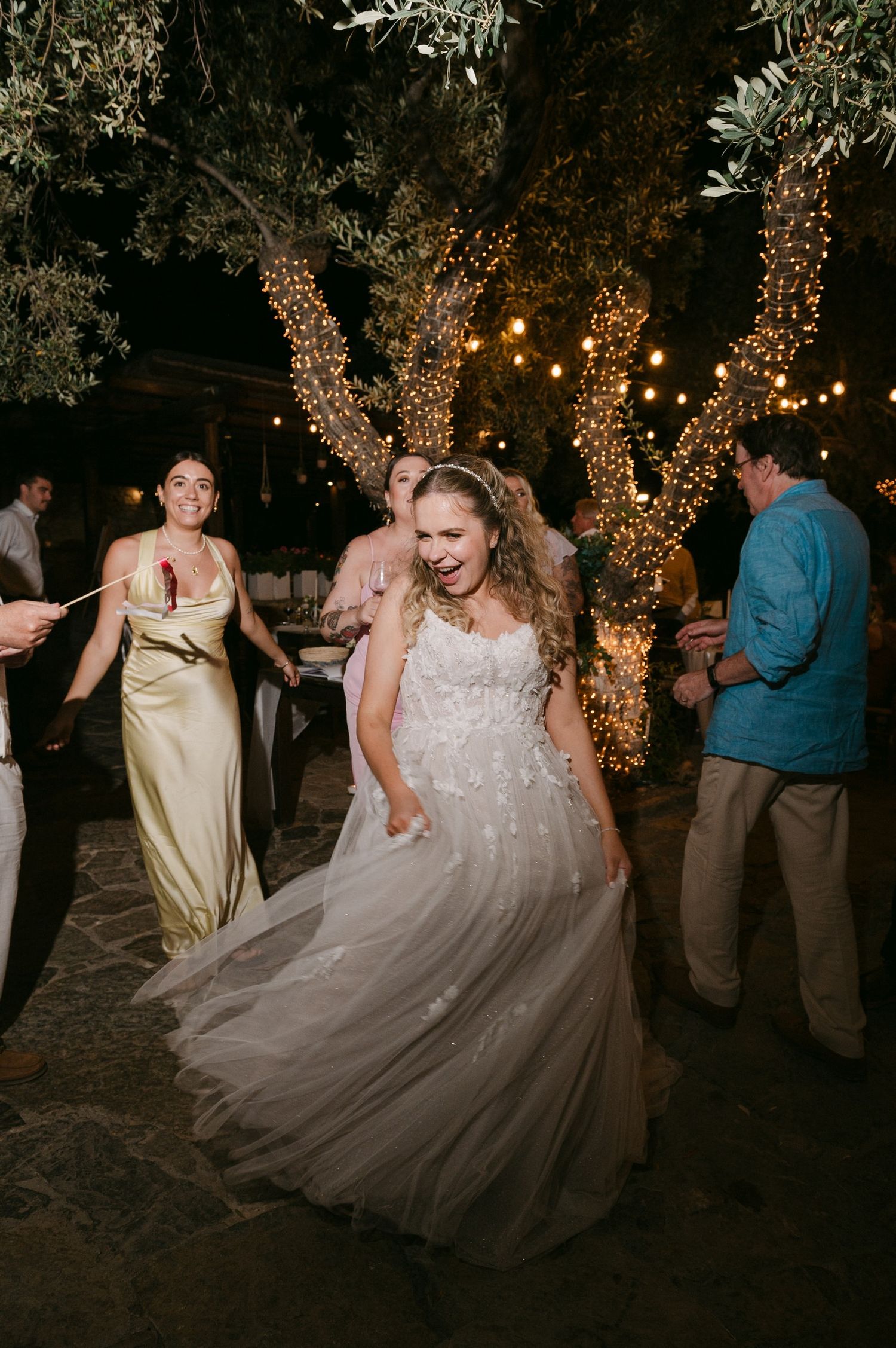 Wedding reception guests dancing and twirling on outdoor dance floor under trees illuminated with string lights at night.